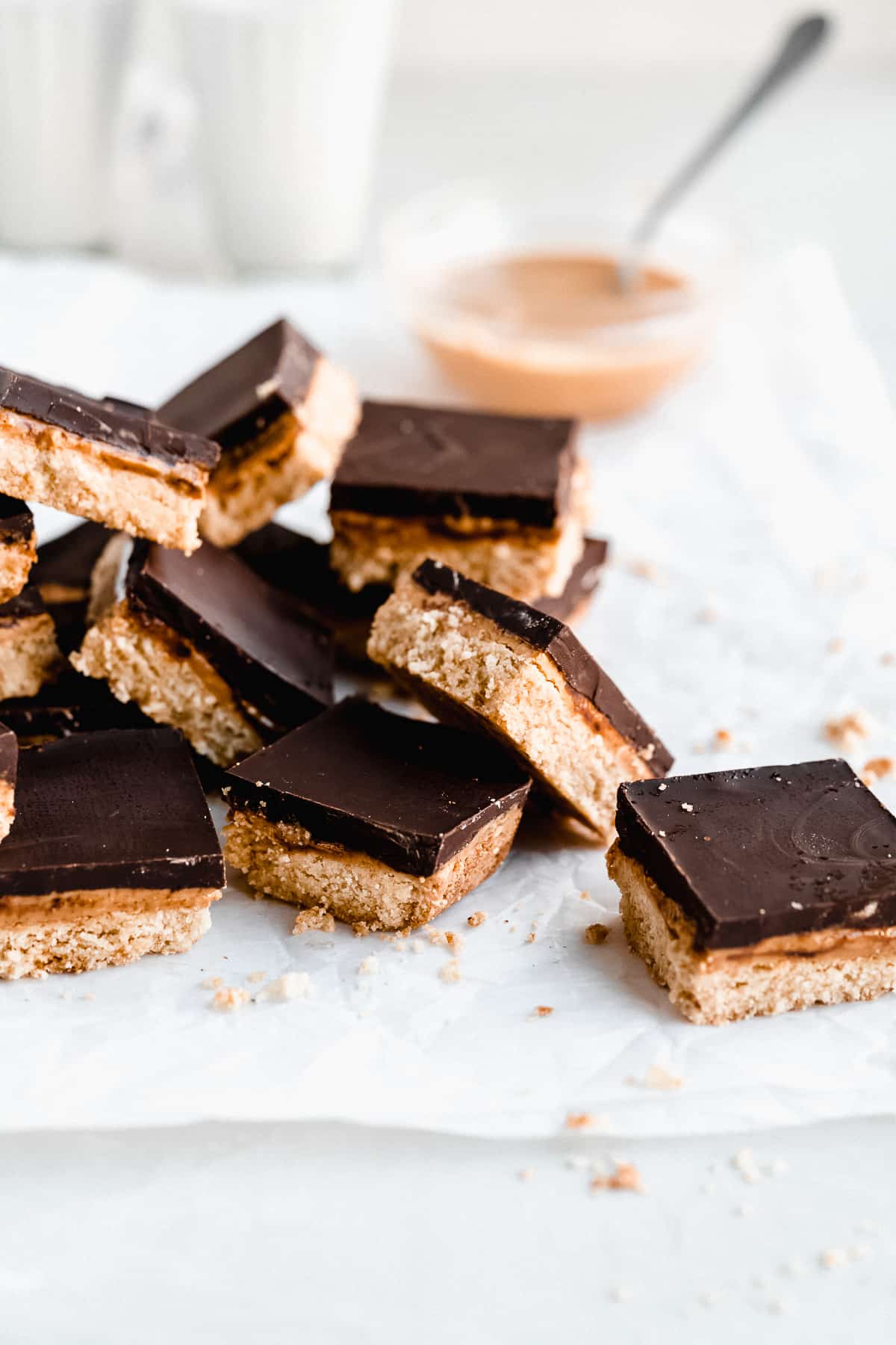 Side view image of several Tagalong Bars cut into squares and mounded on top of one another on parchment paper on a white marble slab.  A small bowl of peanut butter is in the background.  