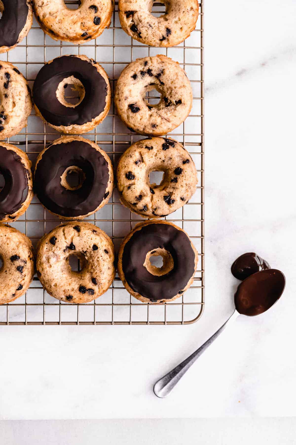 Overhead image of Banana Bread Doughnuts cooling on a wire rack. A spoon with chocolate icing is laying nearby.