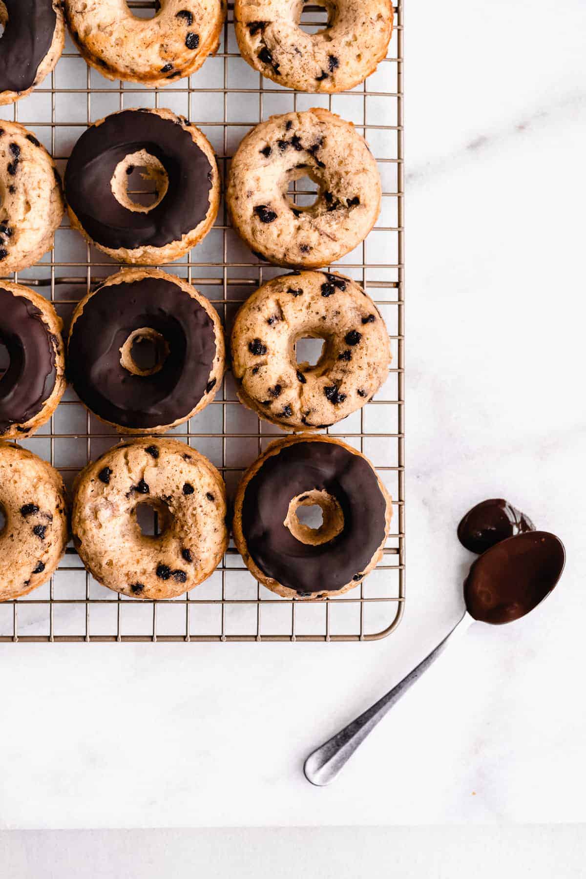 Overhead image of Banana Bread Doughnuts cooling on a wire rack.  A spoon with chocolate icing is laying nearby.  