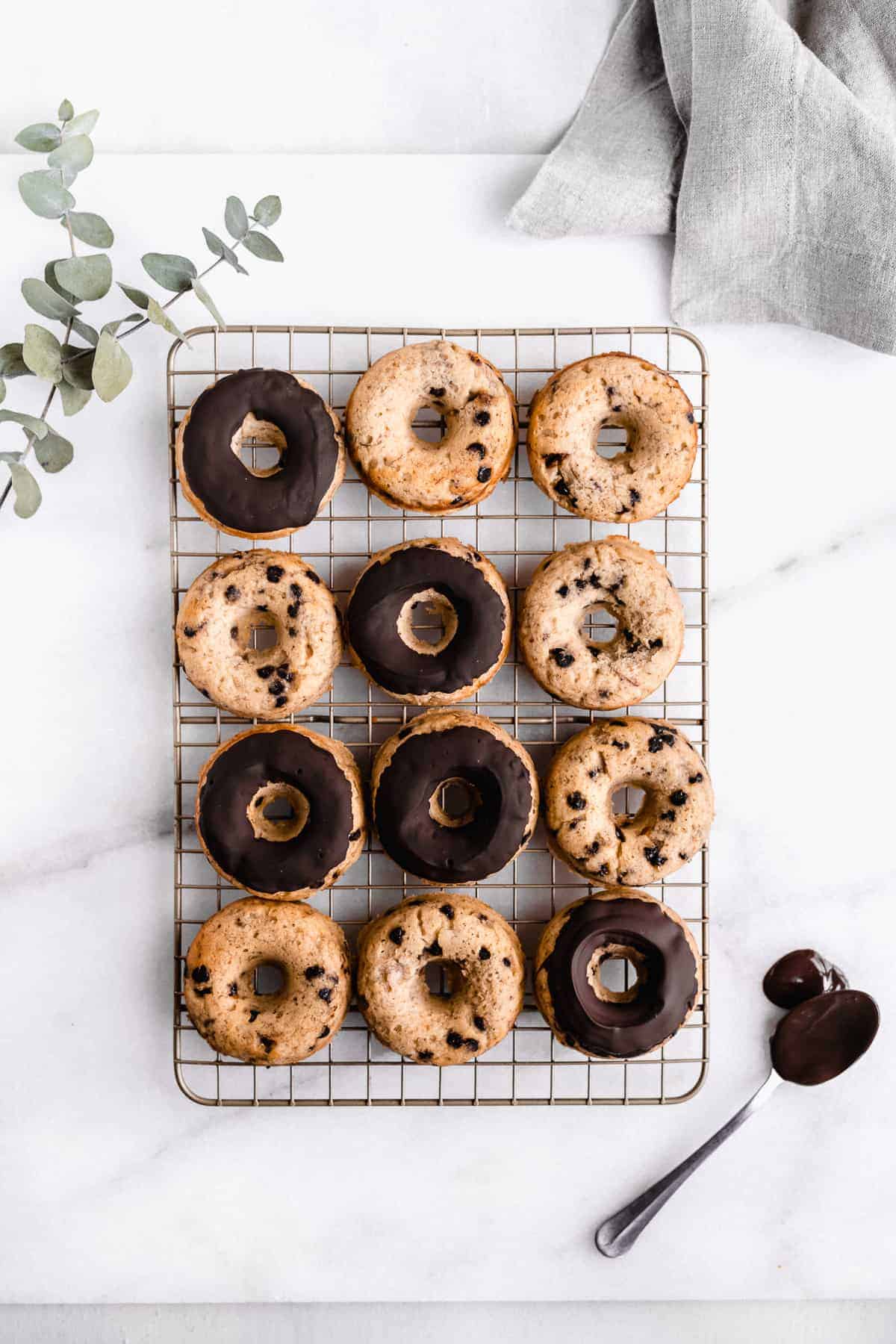 Overhead image of twelve Banana Bread Doughnuts laying on a cooling rack on a white marble slab. Several of them have been frosted with chocolate icing.