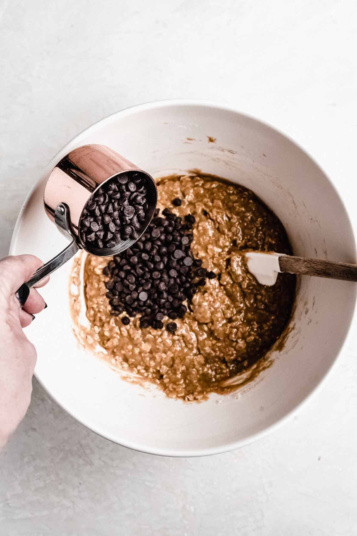 Overhead photo of the process of making this recipe.  A large white bowl has several ingredients in it and a hand is pouring the chocolate chips into the bowl.  