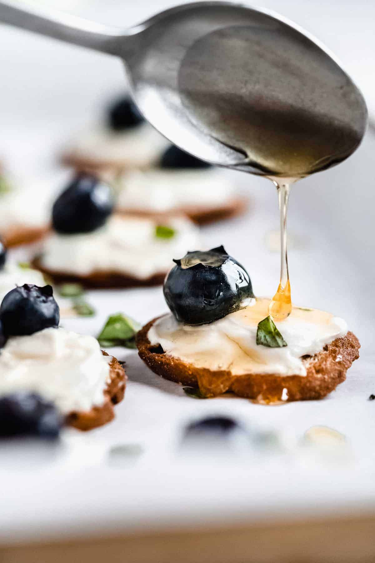 Side view image of a crisp with Honey Whipped Goat Cheese and a blueberry.  A spoon is overhead drizzling honey on top!  