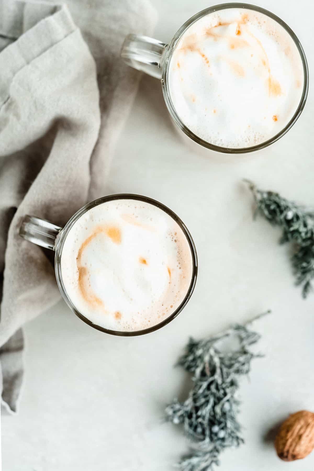 Overhead image of two clear mugs of Maple Sea Salt Almond Milk Lattes sitting on a marble slab with a gray linen cloth nearby.