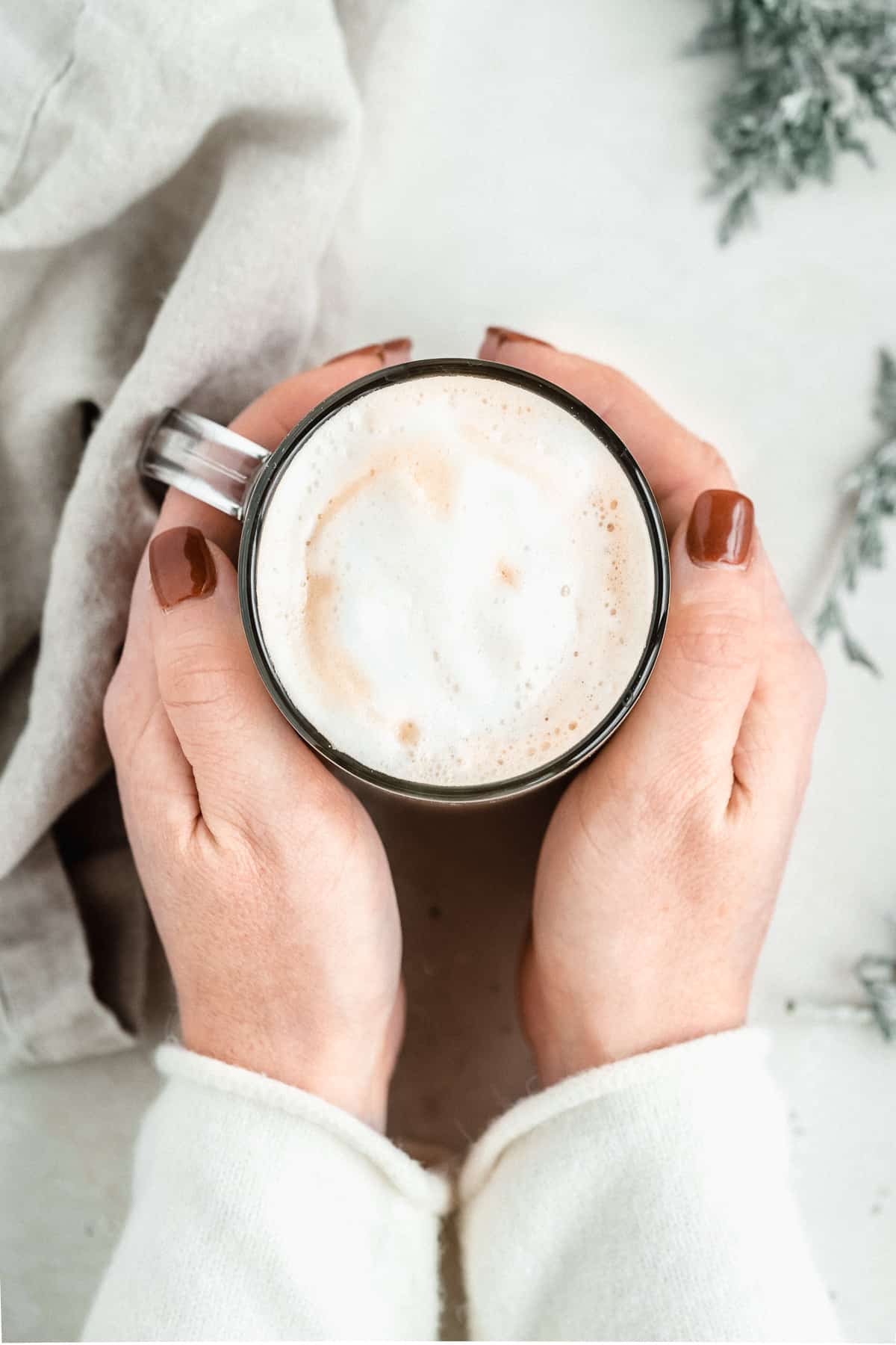 Overhead image of two hands wrapped around a mug of Maple Sea Salt Almond Milk Latte.  A grey linen cloth is laying nearby.  