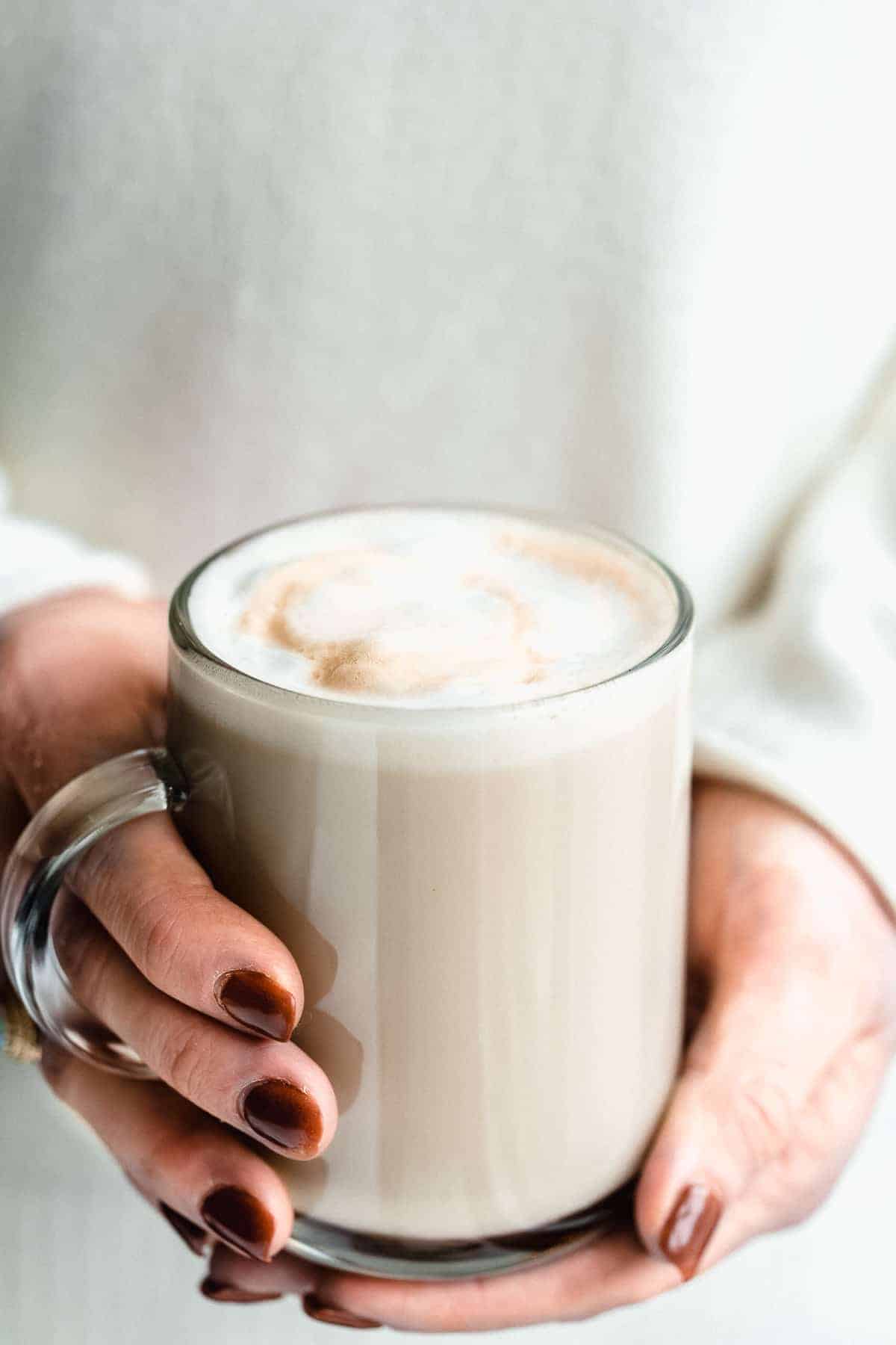 Side view image of two hands holding a clear mug of Maple Sea Salt Almond Milk Latte.  