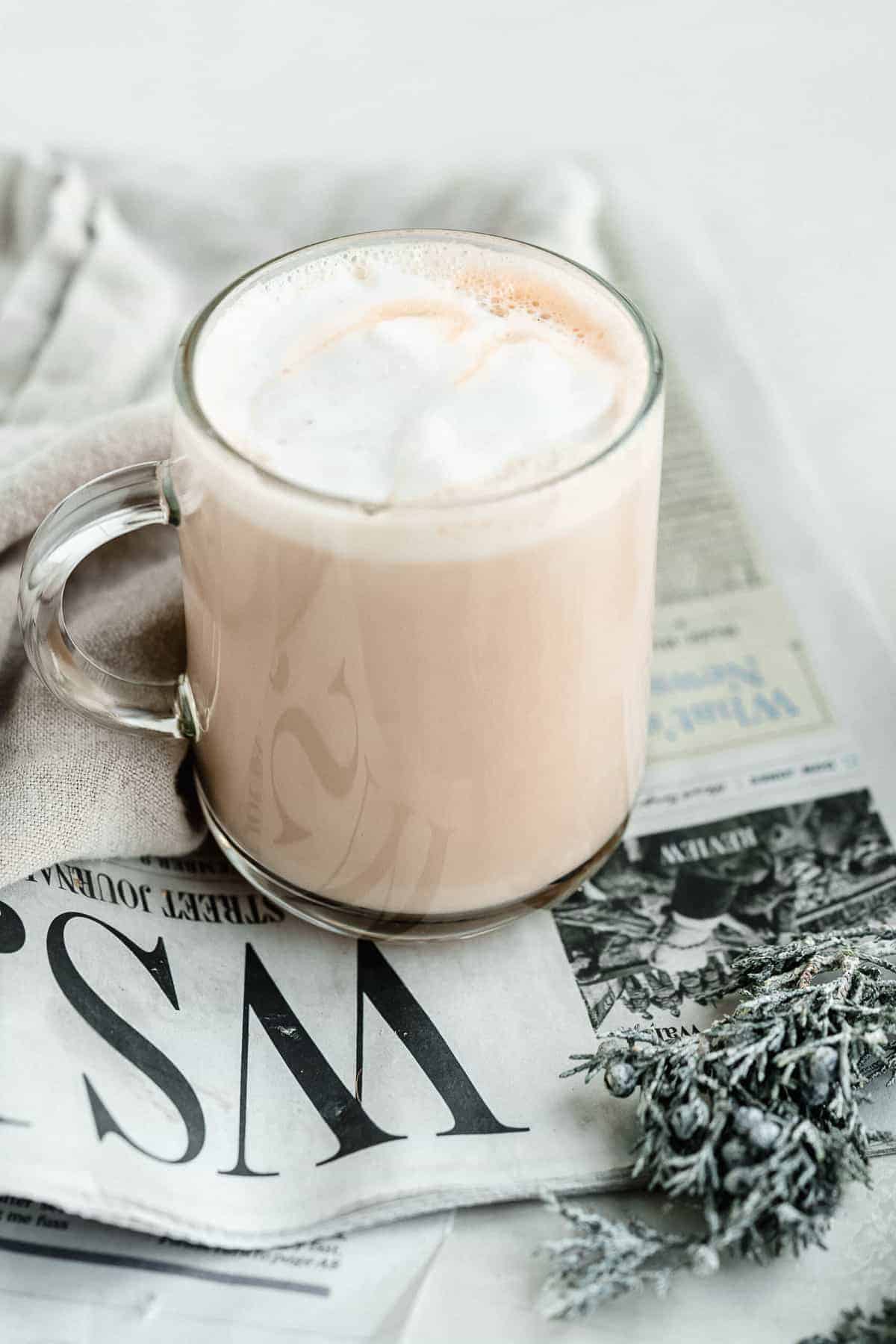 Close up image of a clear mug of Maple Sea Salt Almond Milk Latte sitting on top of a newspaper with a gray linen cloth nearby.  