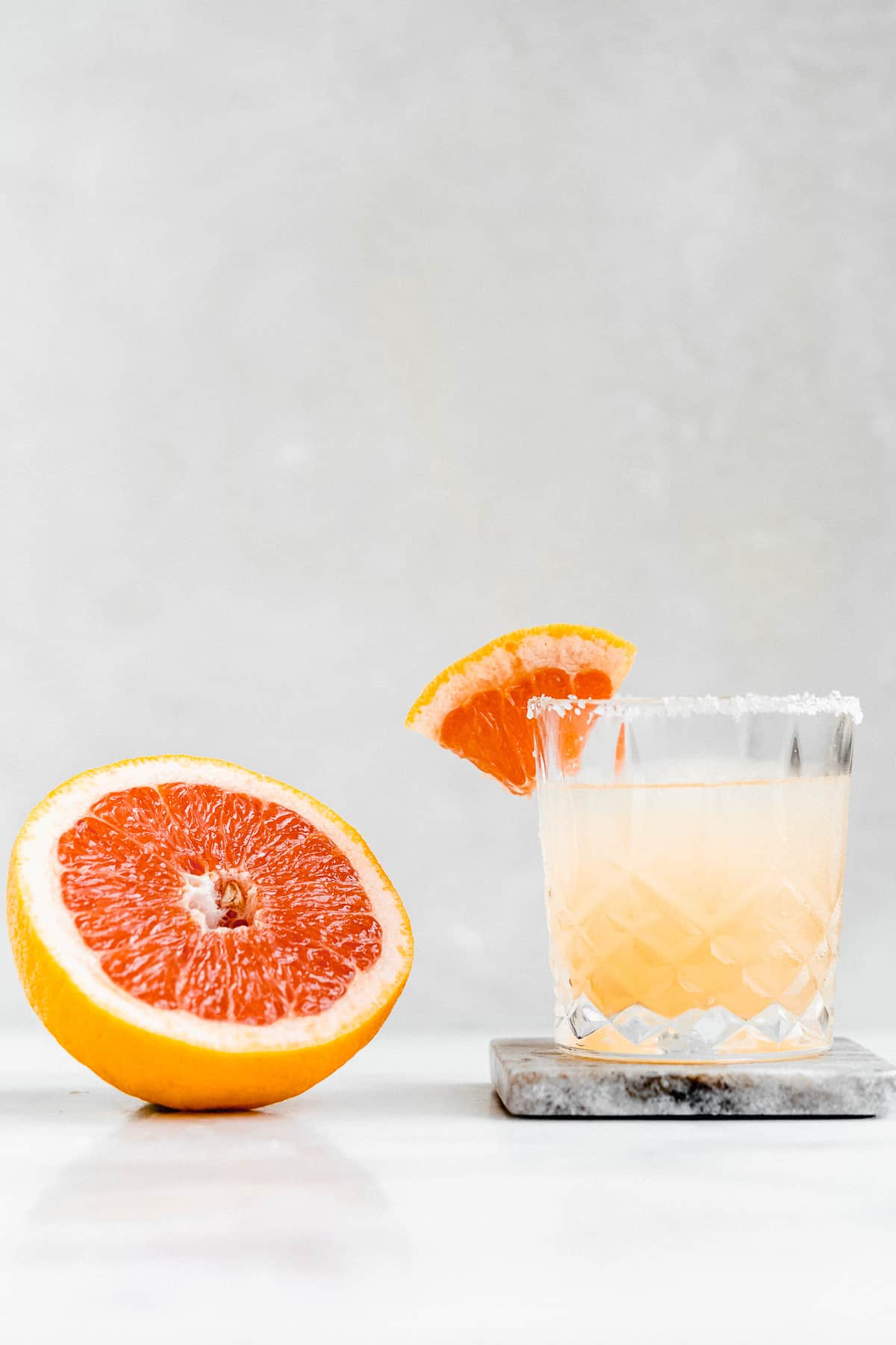 Side view image of a  Skinny Smoky Grapefruit Margarita served in a crystal glass with a salted rim and garnished with a slice of grapefruit.  Half of a grapefruit is laying nearby.  