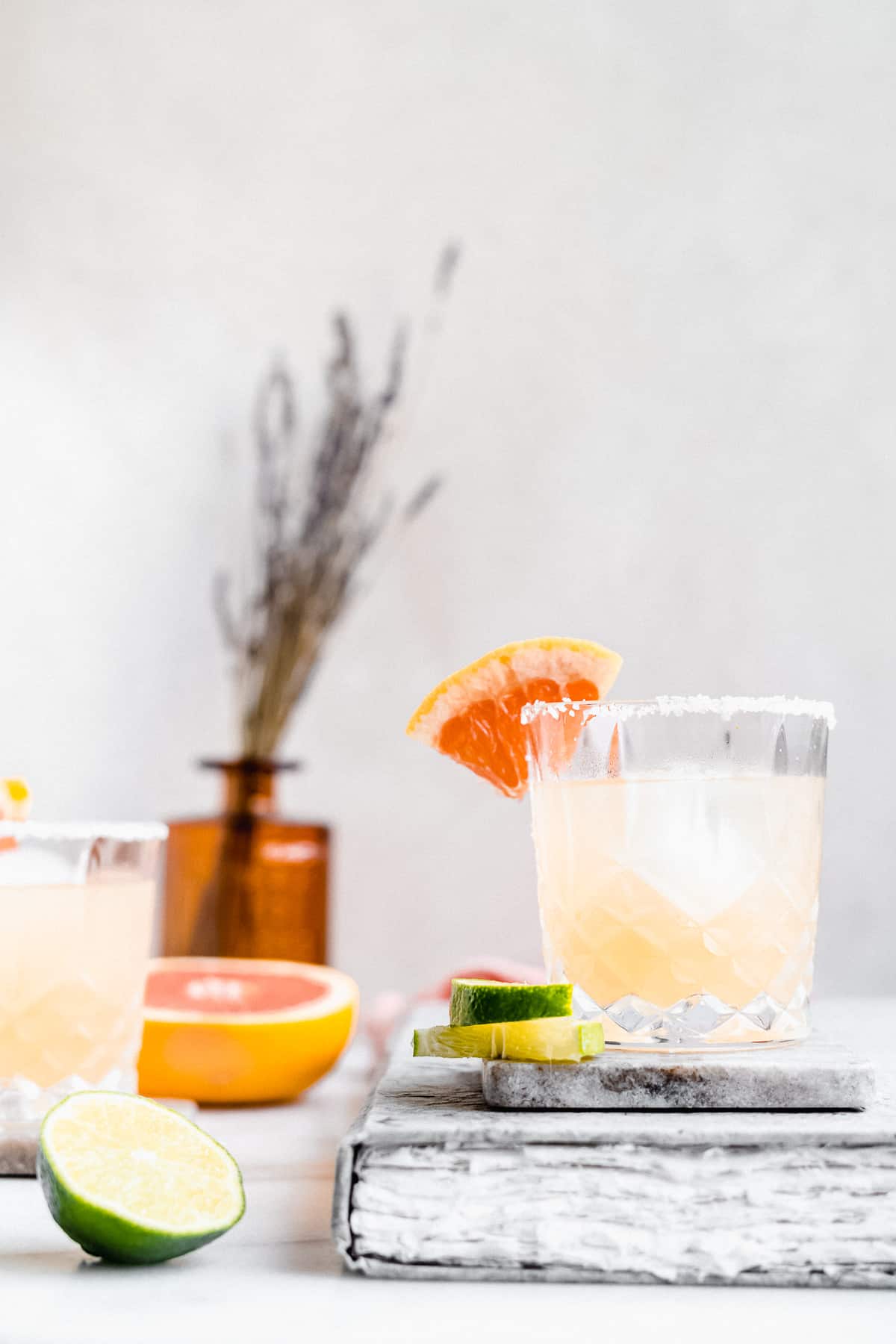 Side view photo of the Skinny Smoky Grapefruit Margarita served in a crystal glass that is sitting on a coaster on top of a book. Half a grapefruit and a slice of lime are seen in the background.