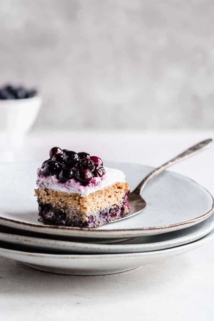 Image of a single serving of the Blueberry Vanilla Sheet Cake being placed on a stack of gray plates with a silver cake server.