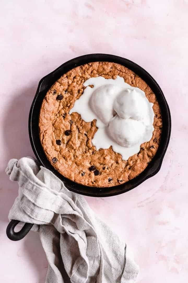 Overhead image of freshly baked Paleo Chocolate Chip Cookie Skillet served a la mode with vanilla ice cream melting on top. A gray linen towel is wrapped around the handle of the cast iron skillet, which is sitting on a pink background.