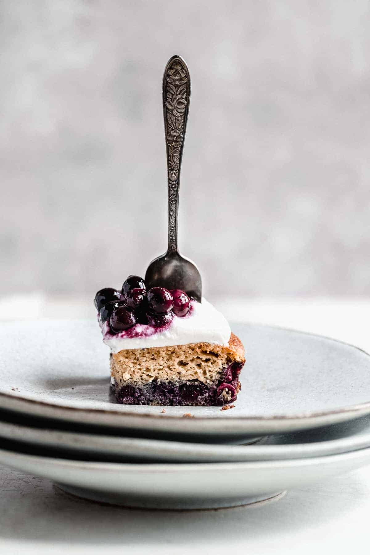 Side view photo of a single serving of Blueberry Vanilla Sheet Cake on a stack of gray plates with a silver fork sticking up from the cake.  