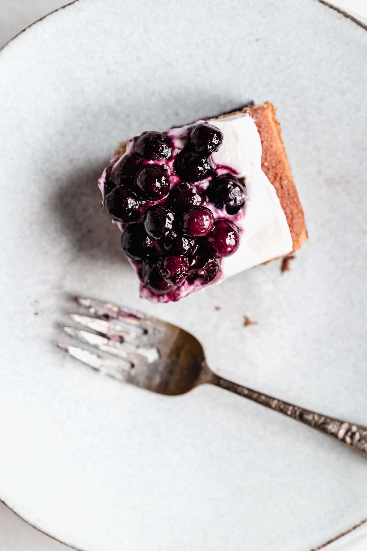 Overhead photo of a single serving of Blueberry Vanilla Sheet Cake on a gray plate with a silver fork laying nearby.