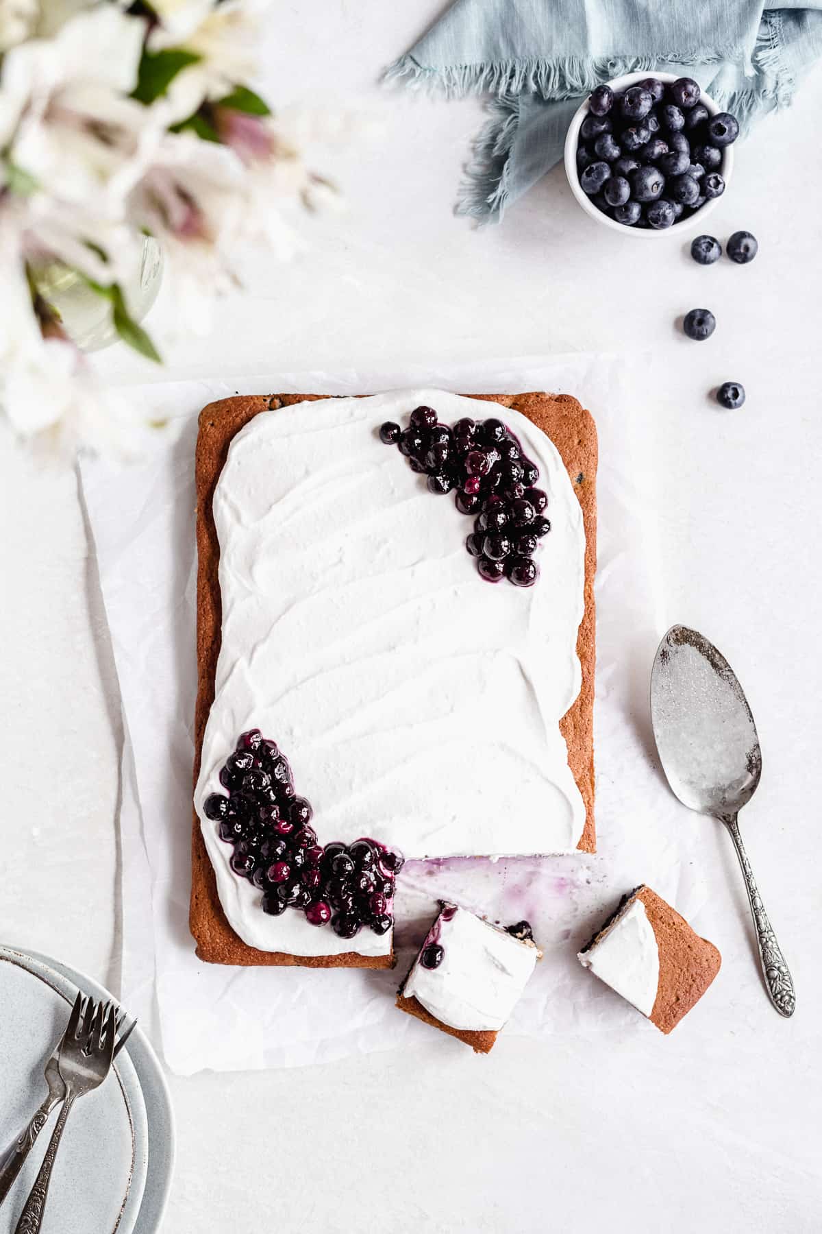 Overhead photo of the whole Blueberry Vanilla Sheet Cake on a white marble slab with two pieces cut into squares with a silver cake server and pulled away. Several small plates and forks are placed nearby.