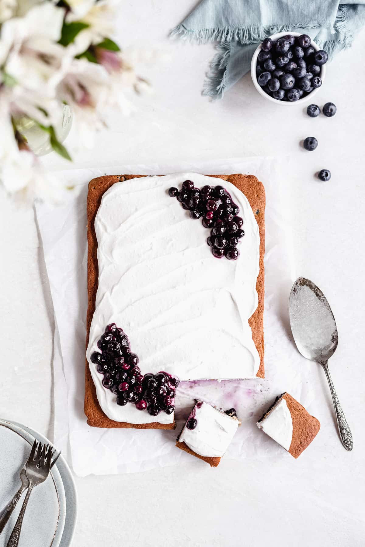 Overhead photo of the whole Blueberry Vanilla Sheet Cake on a white marble slab with two pieces cut into squares with a silver cake server and pulled away.  Several small plates and forks are placed nearby.  