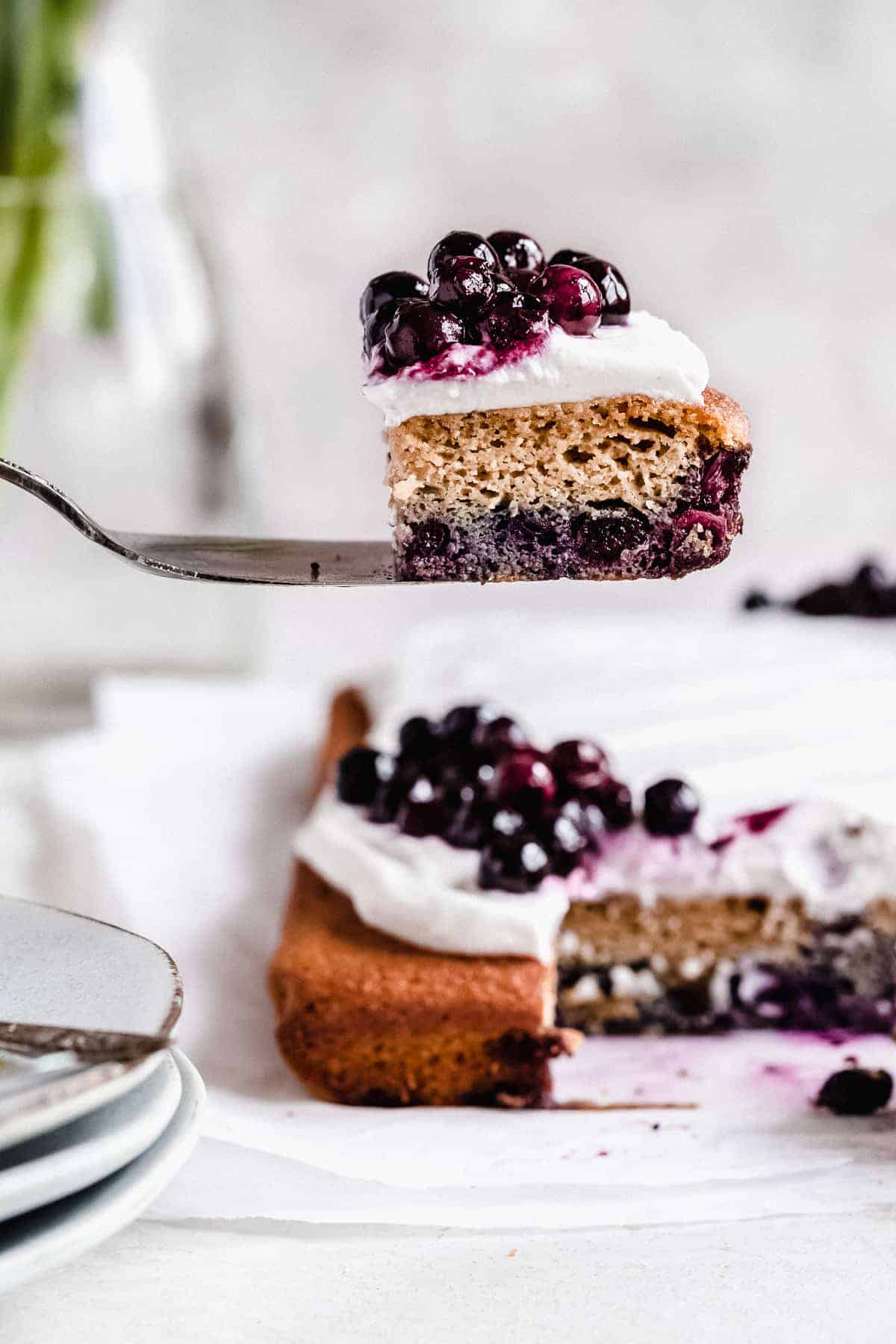 Side view photo of a piece of Blueberry Vanilla Sheet Cake sitting on a silver cake server in the air above the sheet cake. Several small plates and forks can be seen below.