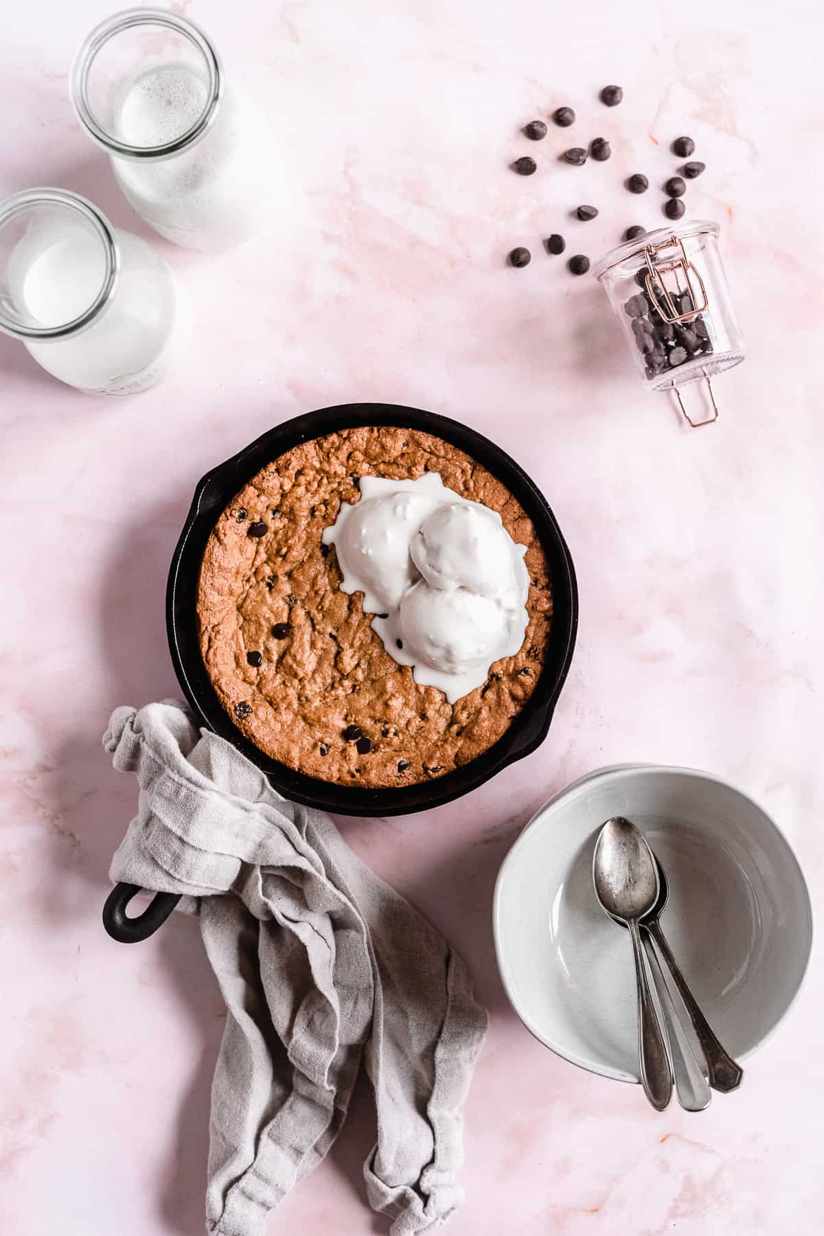 Overhead image of a Paleo Chocolate Chip Cookie Skillet in a cast iron skillet placed on a pink background.  A gray linen towel is wrapped around the handle of the skillet.  Several gray bowls and spoons are placed nearby.  Two glasses of milk can be seen in the background and chocolate chips are scattered around.  