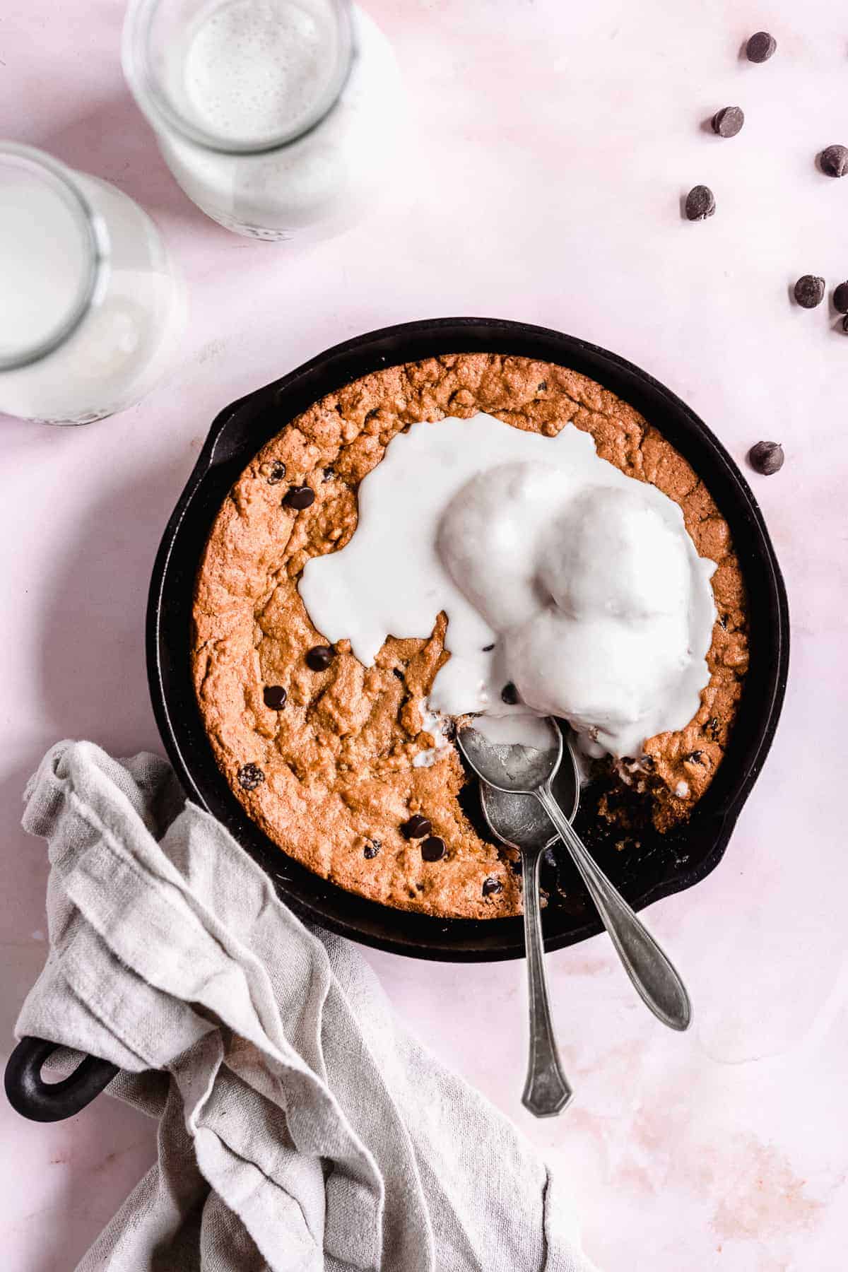 Overhead photo of Paleo Chocolate Chip Cookie Skillet in a cast iron skillet served a la mode with vanilla ice cream sitting on a pink background. Two glasses of milk are sitting nearby. A serving has been removed and two spoons are placed in the skillet.