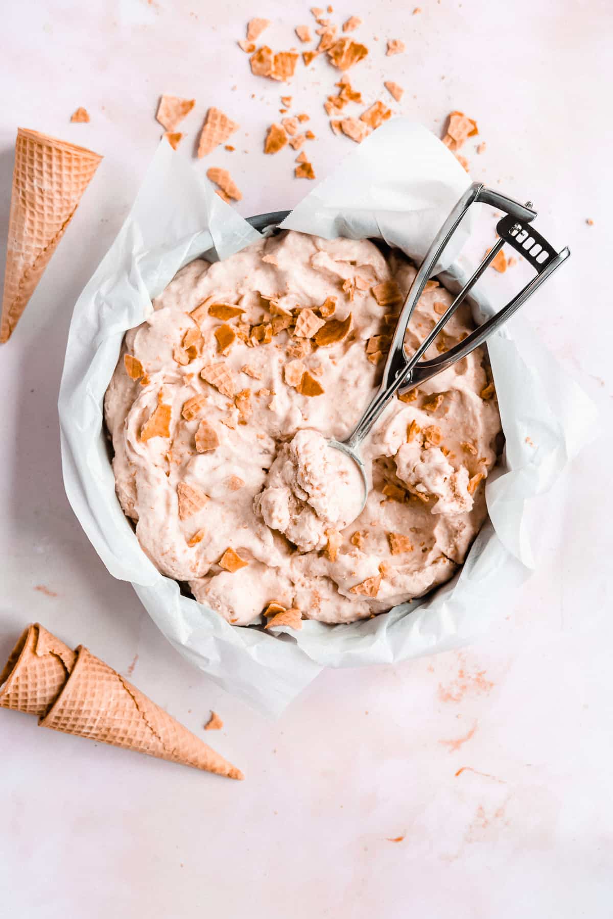 Overhead image of Homemade Vegan Peach Ice Cream on a marble slab with an ice cream scoop filled with delicious ice cream.  Several  cones are laying nearby.  