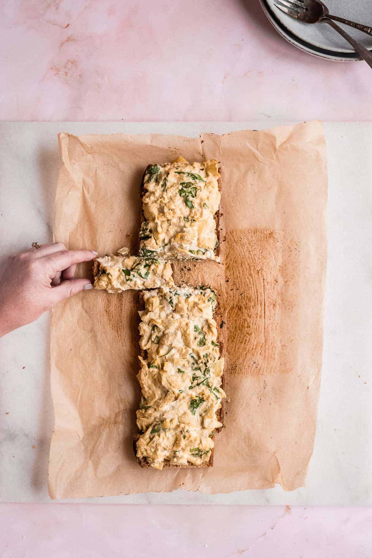 Overhead image of the full loaf of Spinach Artichoke Toast laying on parchment paper. A single stick has been cut and a hand is pulling the stick away from the loaf.