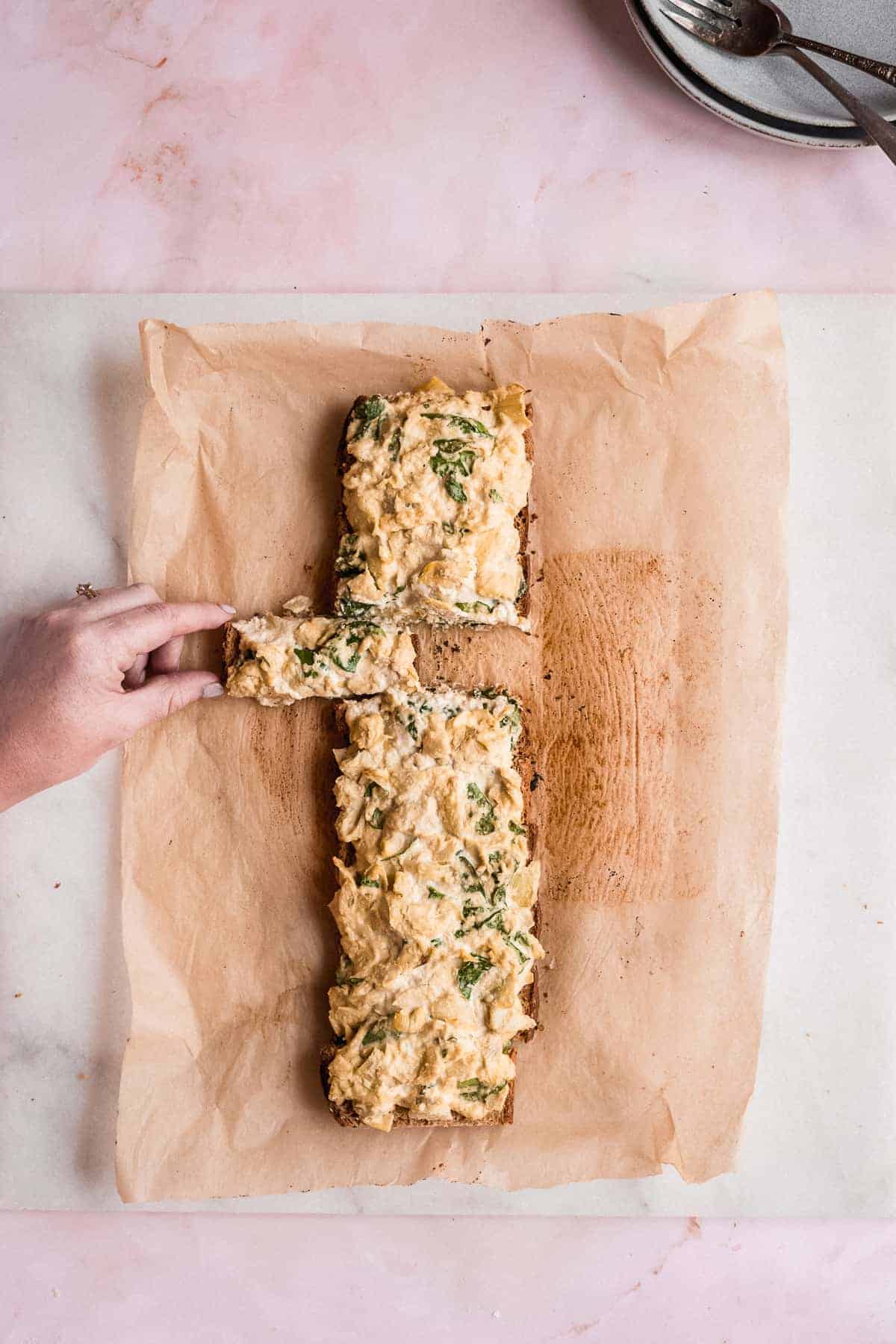 Overhead image of the full loaf of Spinach Artichoke Toast laying on parchment paper.  A single stick has been cut and a hand is pulling the stick away from the loaf.  