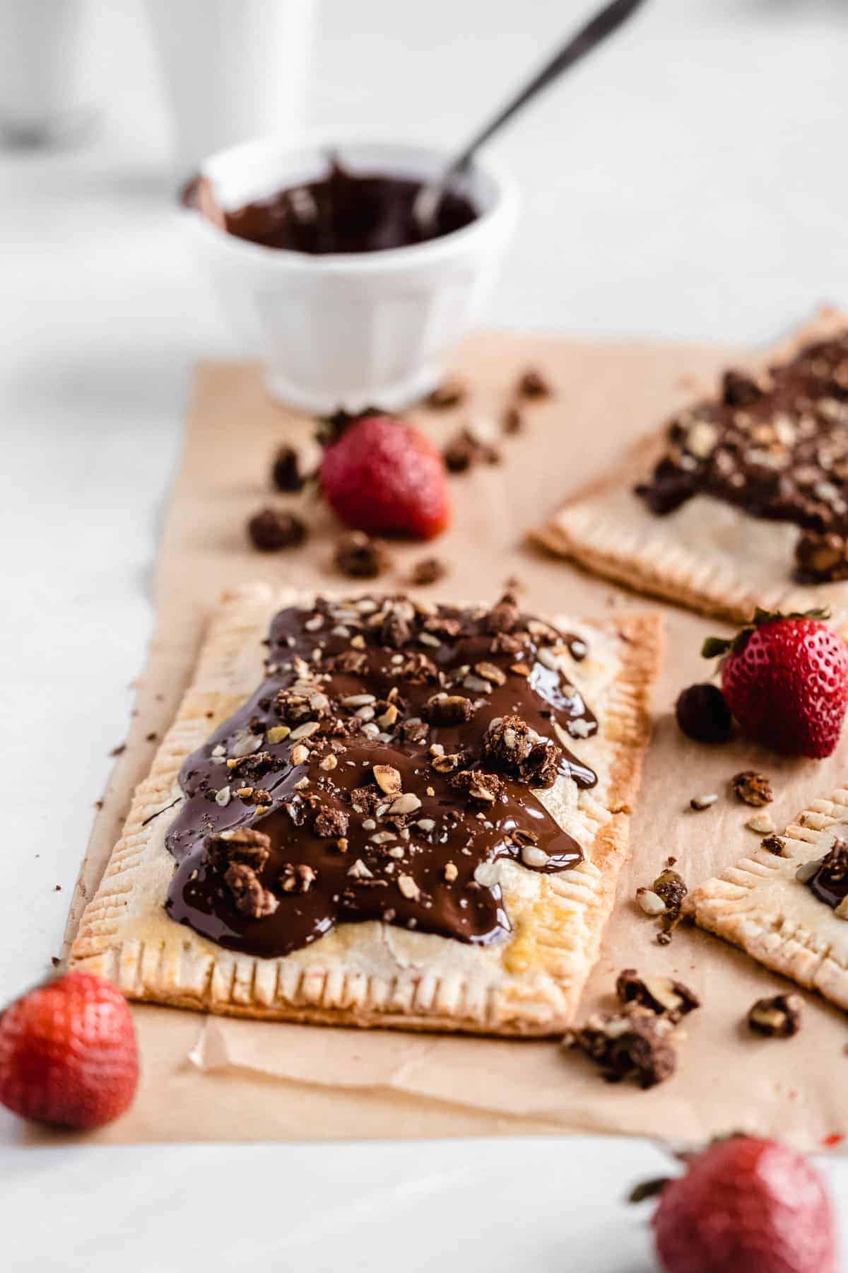 Side view photo of completed Chocolate Covered Strawberry Pop Tarts arranged on parchment paper. A small white bowl of melted chocolate sauce is in the background. Several strawberries are scattered around.