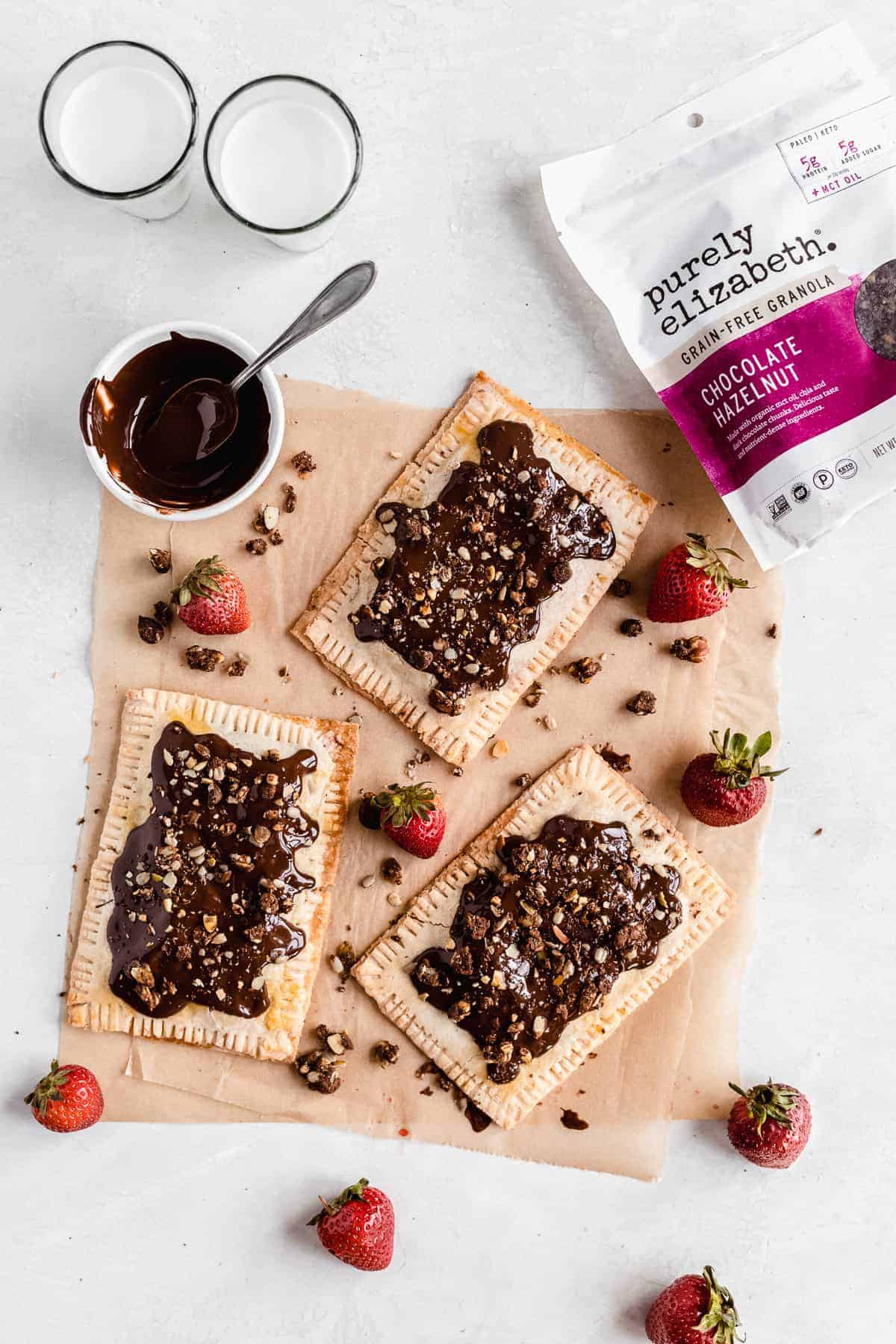 Overhead photo of three completed tarts arranged on parchment paper with several strawberries scattered around.  A bag of Purely Elizabeth granola and a small bowl of chocolate sauce can be seen at the top of the photo.  