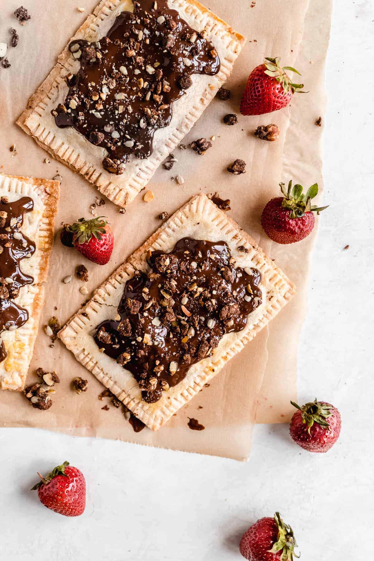 Overhead photo of completed Chocolate Covered Strawberry Pop Tarts arranged on parchment paper on a marble slab.  Several strawberries are scattered nearby.  