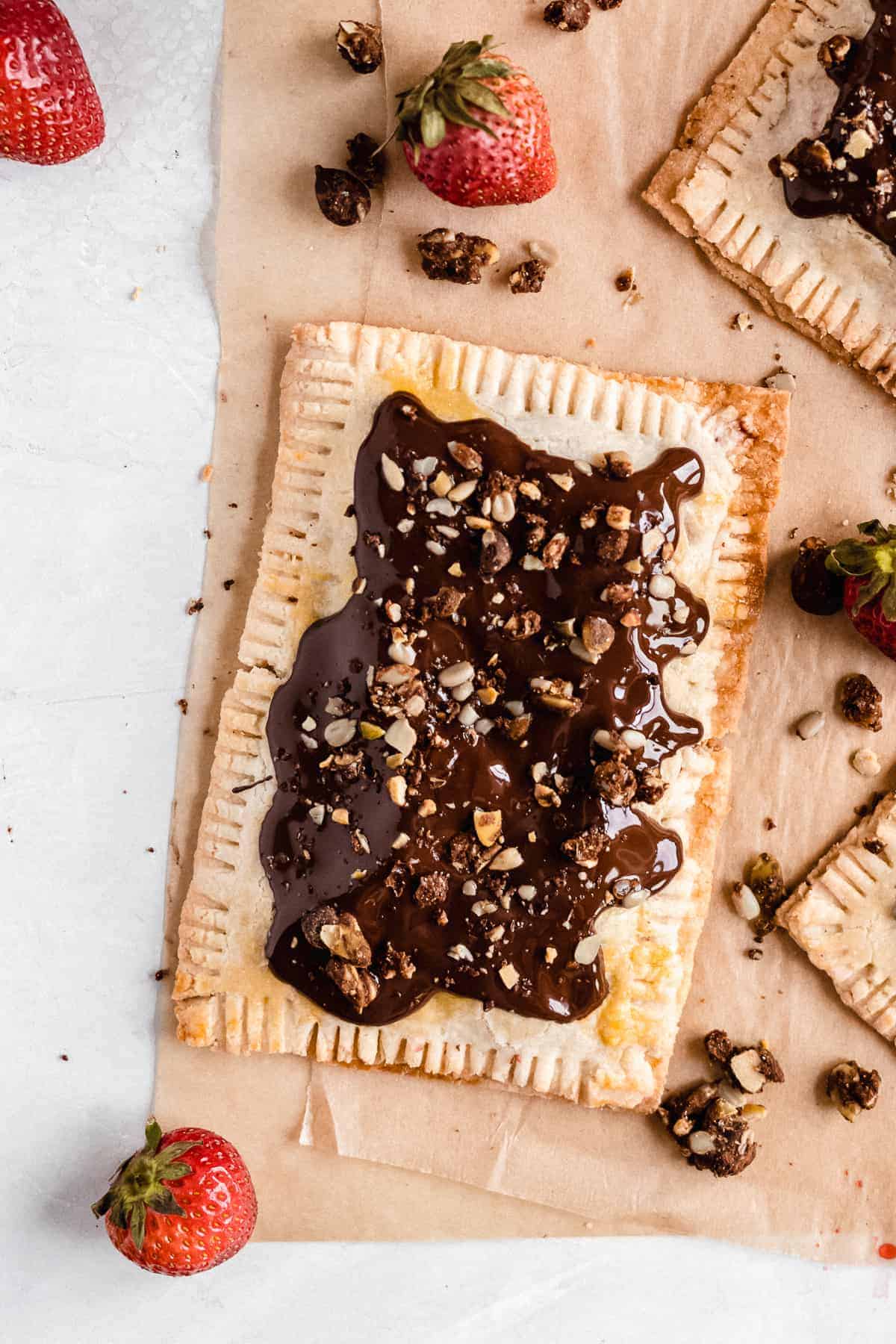 Overhead photo of a single Chocolate Covered Strawberry Pop Tart on parchment paper with several strawberries sprinkled around.
