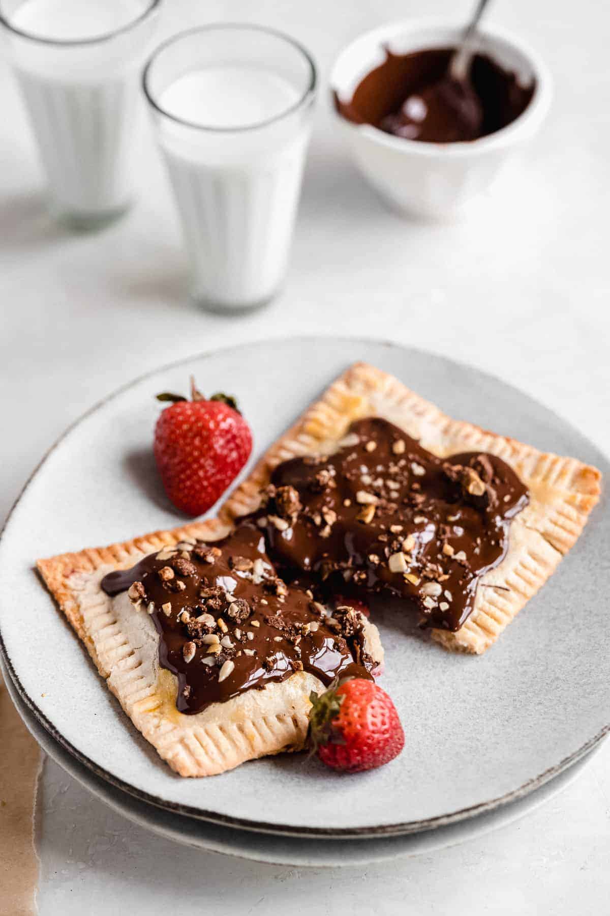 Image of a single Chocolate Covered Strawberry Pop Tart on a gray plate with two strawberries placed nearby. Two glasses of milk and a small white bowl of extra chocolate sauce can be seen in the background.
