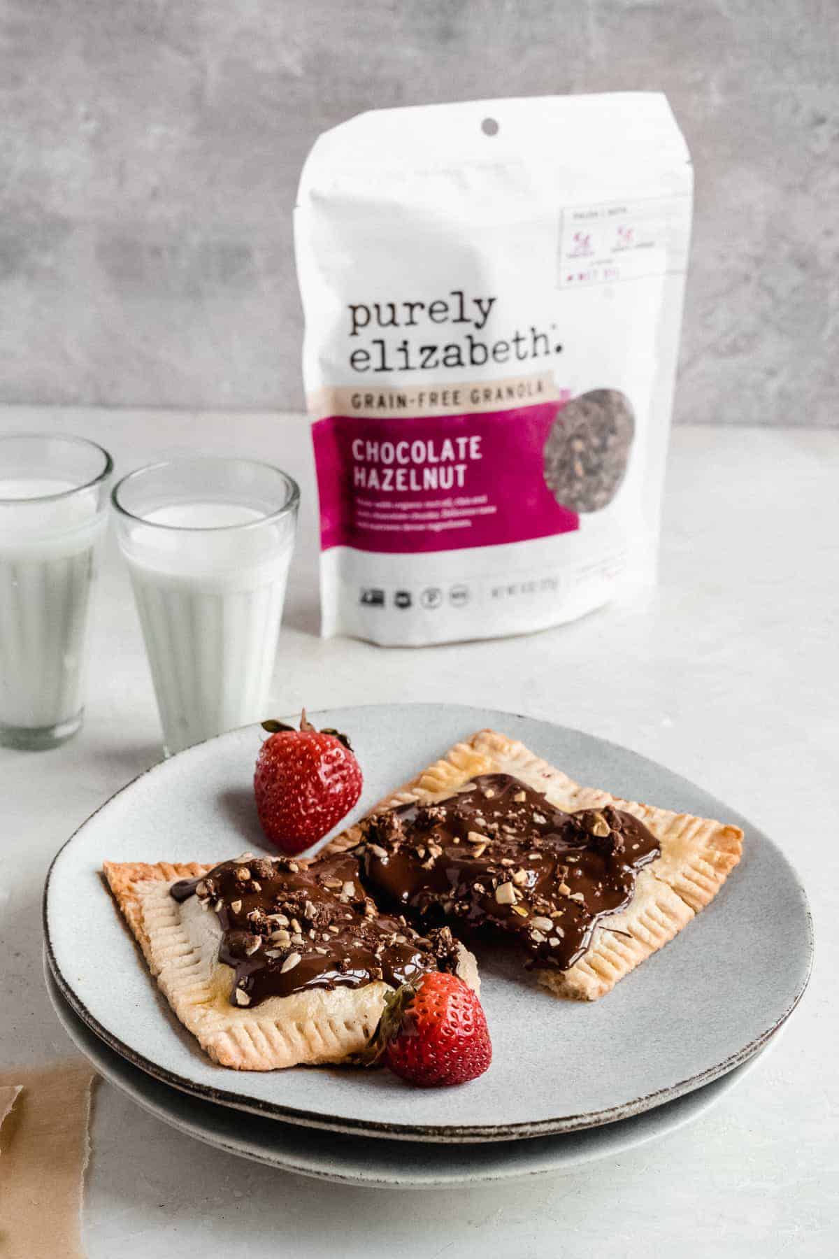 Image of a single Chocolate Covered Strawberry Pop Tart on a gray plate. Two glasses of milk and a bag of Purely Elizabeth granola are in the background.