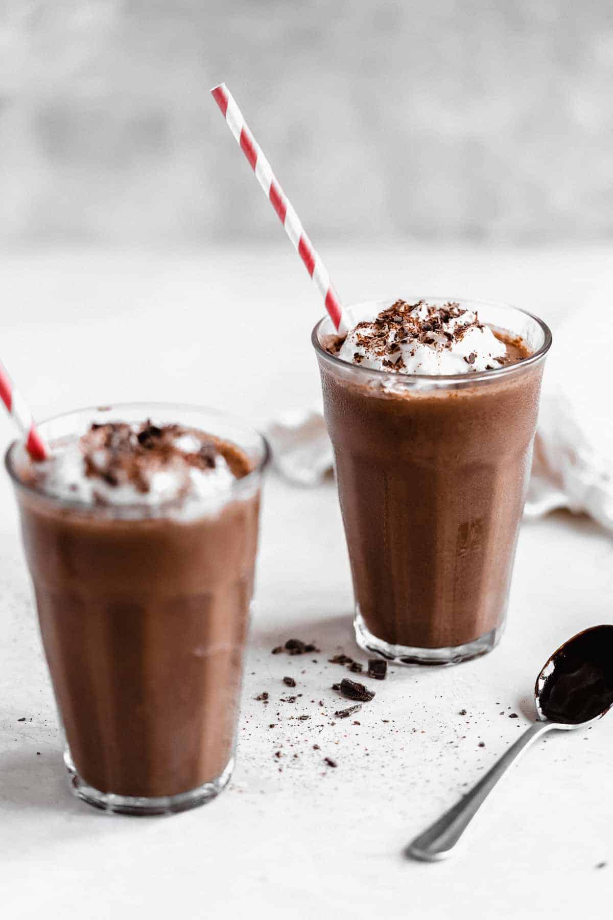 Two glasses of Frozen Mexican Hot Chocolate on a marble slab with red and white striped straws. A spoon and white linen cloth are in the background.