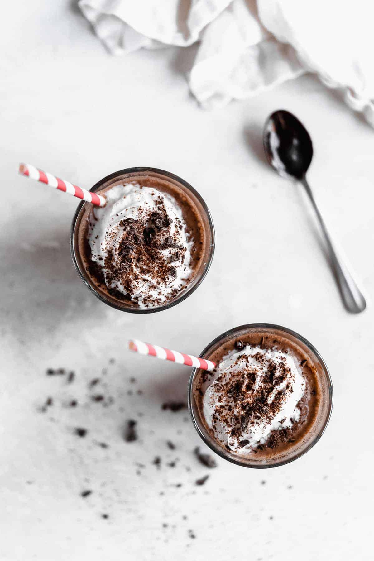 Overhead photo of two glasses of Frozen Mexican Hot Chocolate arranged on a white marble slab.  A silver spoon and white linen cloth can be seen in the background.  