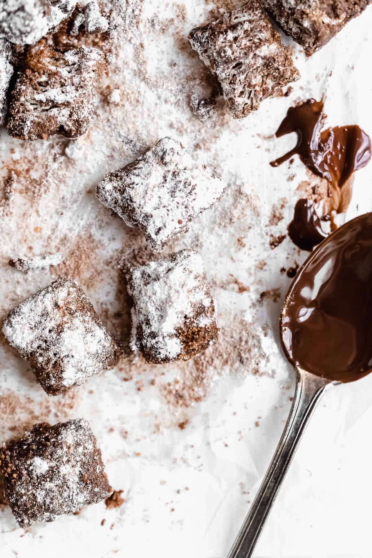 Closeup photo of Healthy Puppy Chow pieces on white parchment paper.  A silver spoon with melted chocolate sauce is laying nearby.  
