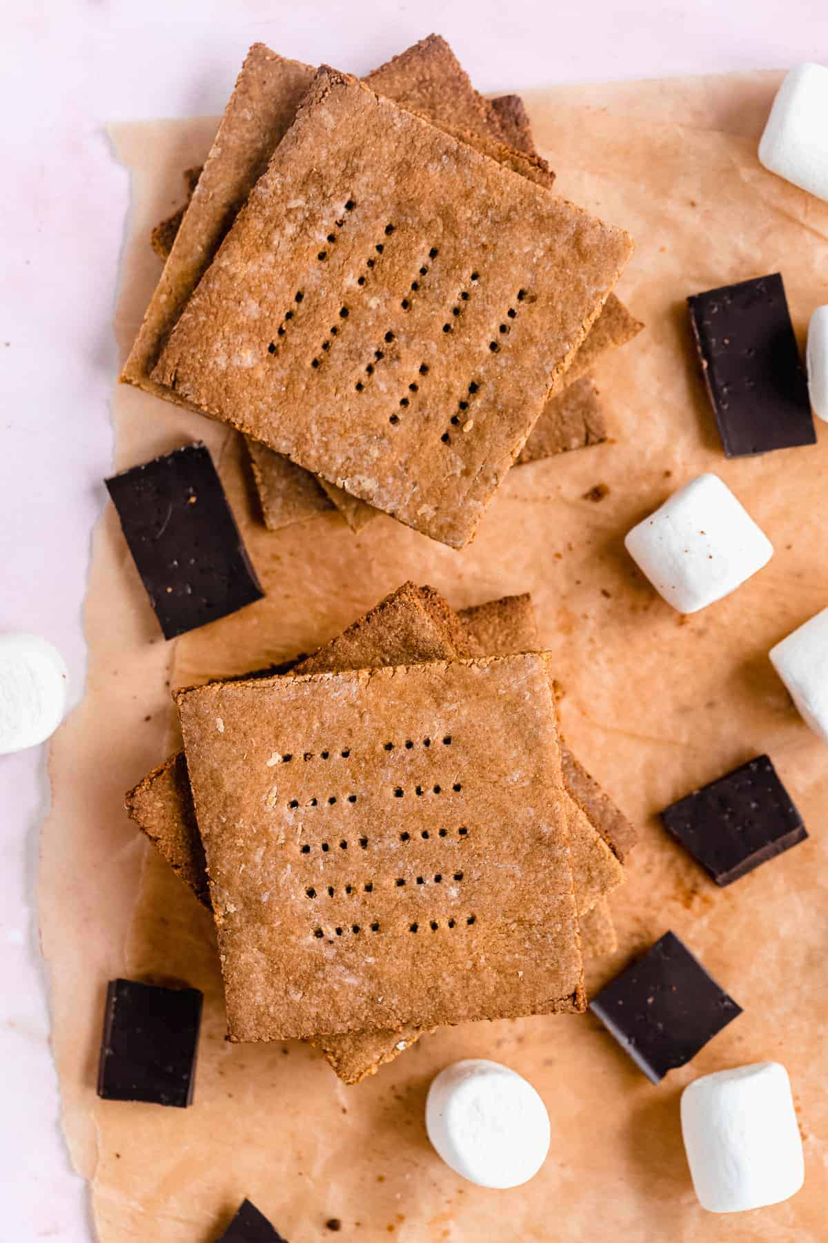 Close up photo of Homemade Graham Crackers stacked on top of one another on parchment paper.  Chocolate pieces and marshmallows are scattered around.  