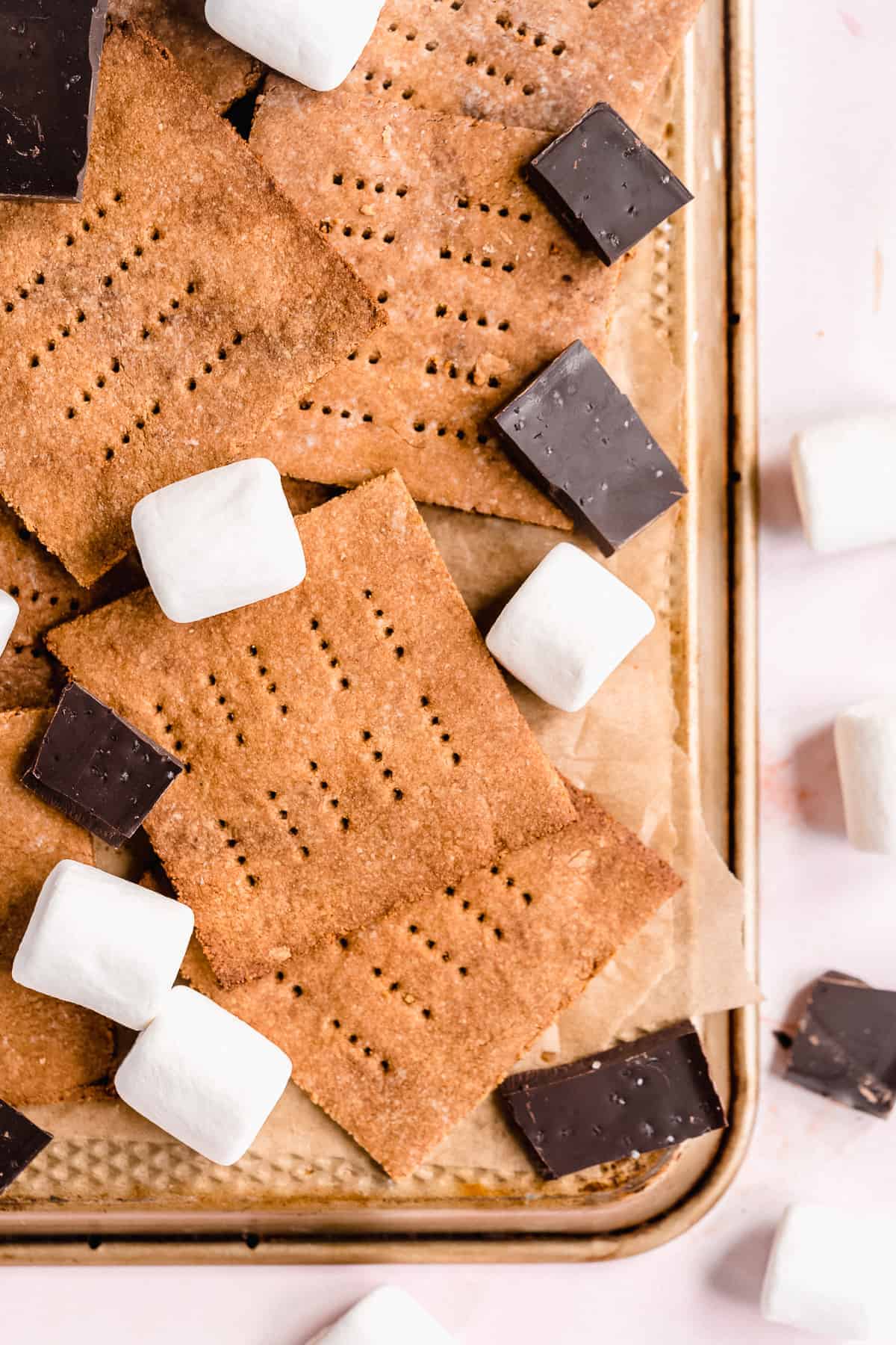 Overhead photo of the completed Homemade Graham Crackers arranged on parchment paper on a baking tray.  Chocolate pieces and marshmallow are scattered around.  