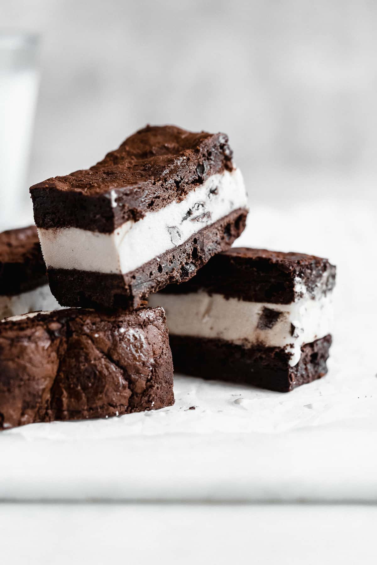 Several Brownie Ice Cream Sandwich squares stacked on top of one another on a white marble slab.  A glass of milk can be seen in the background.  