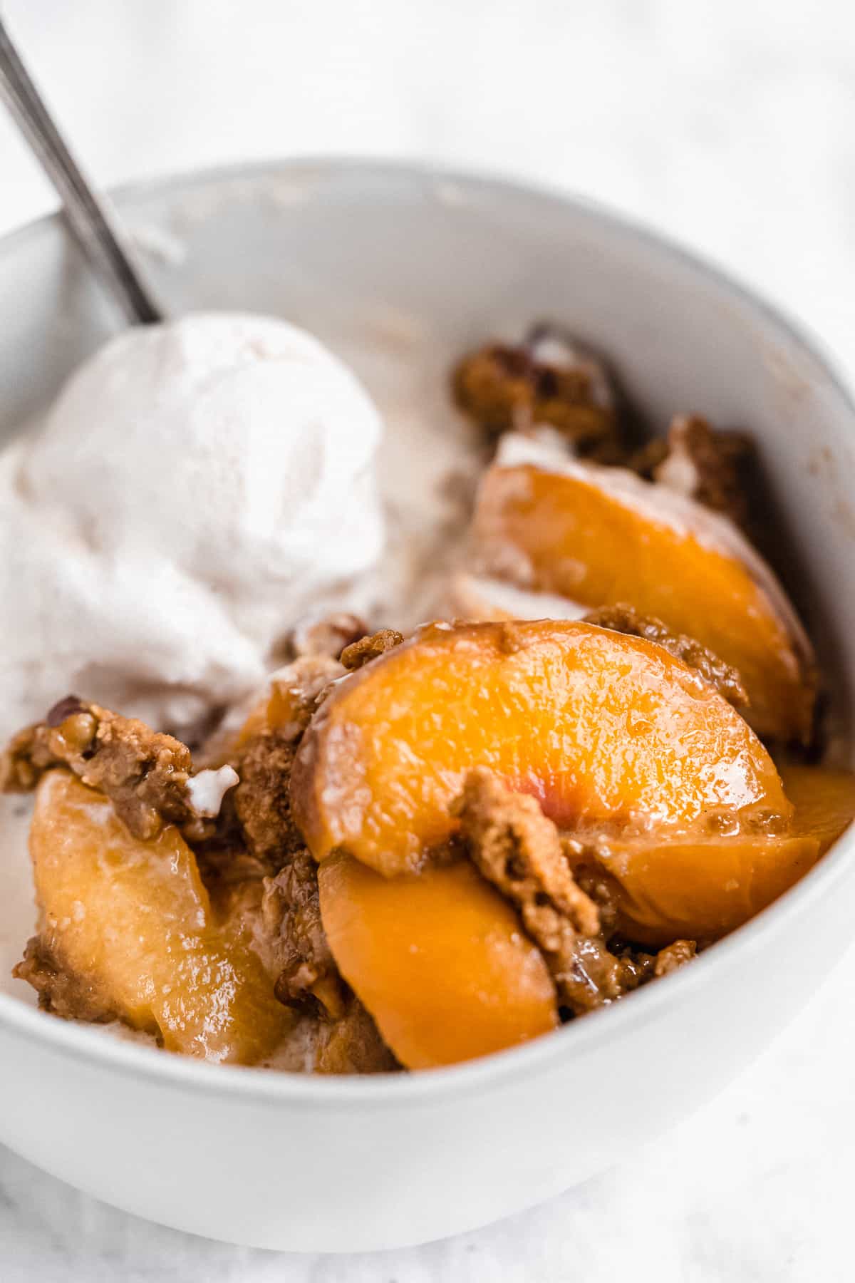 Side view closeup of a serving of Gluten-free Peach Cobbler in a white bowl with a scoop of vanilla ice cream.  