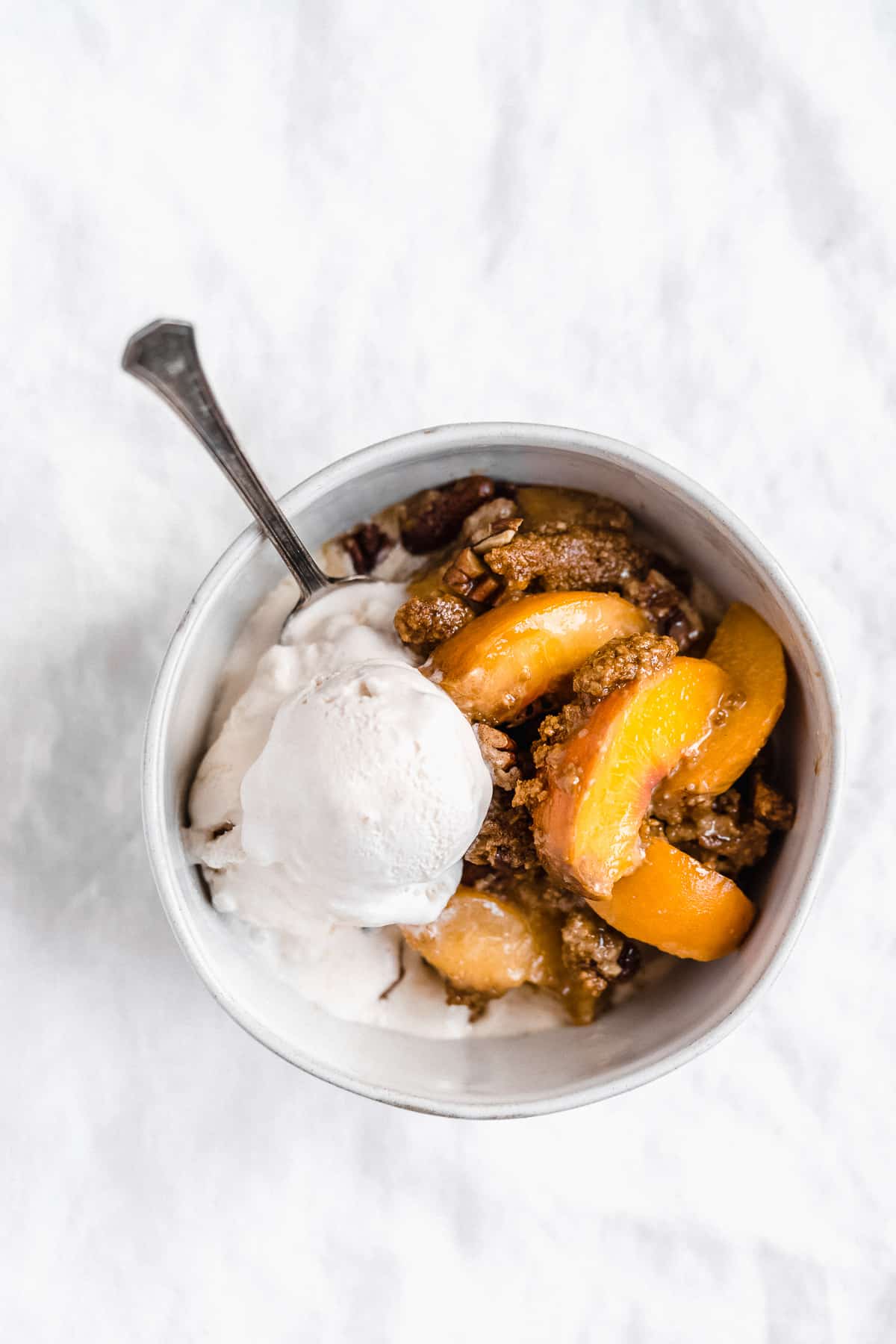 Overhead photo of the freshly baked Gluten-free Peach Cobbler Skillet served in a white bowl and topped with a scoop of vanilla ice cream.  A spoon is in the bowl.  