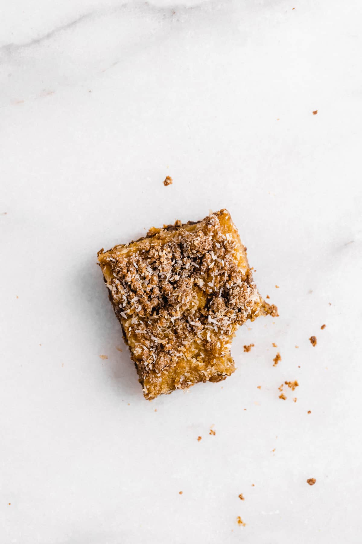 Overhead photo of a single Pineapple Coconut Crumb Bar laying on a white marble slab.  A bite has been taken out of it.  