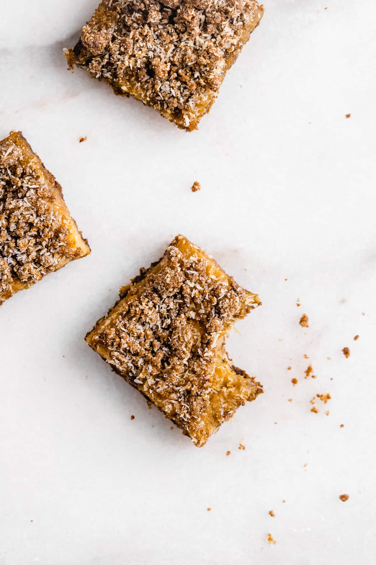 Several squares of Pineapple Coconut Crumb Bars arranged on a white marble slab.  One square has a bite taken out of it.  