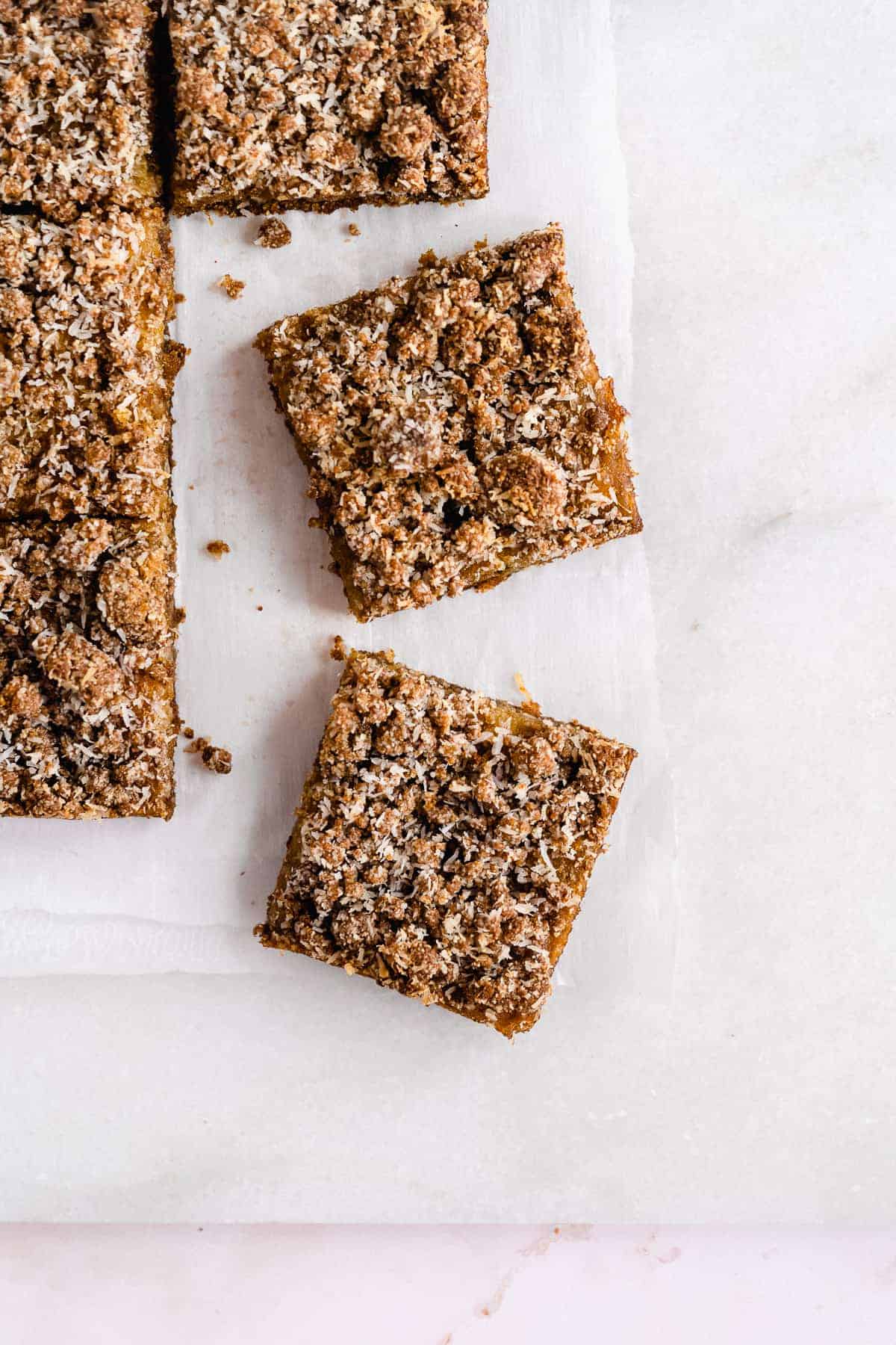 Overhead photo of the completed Pineapple Coconut Crumb Bars that have been cut into squares and arranged on white parchment paper on a marble slab.