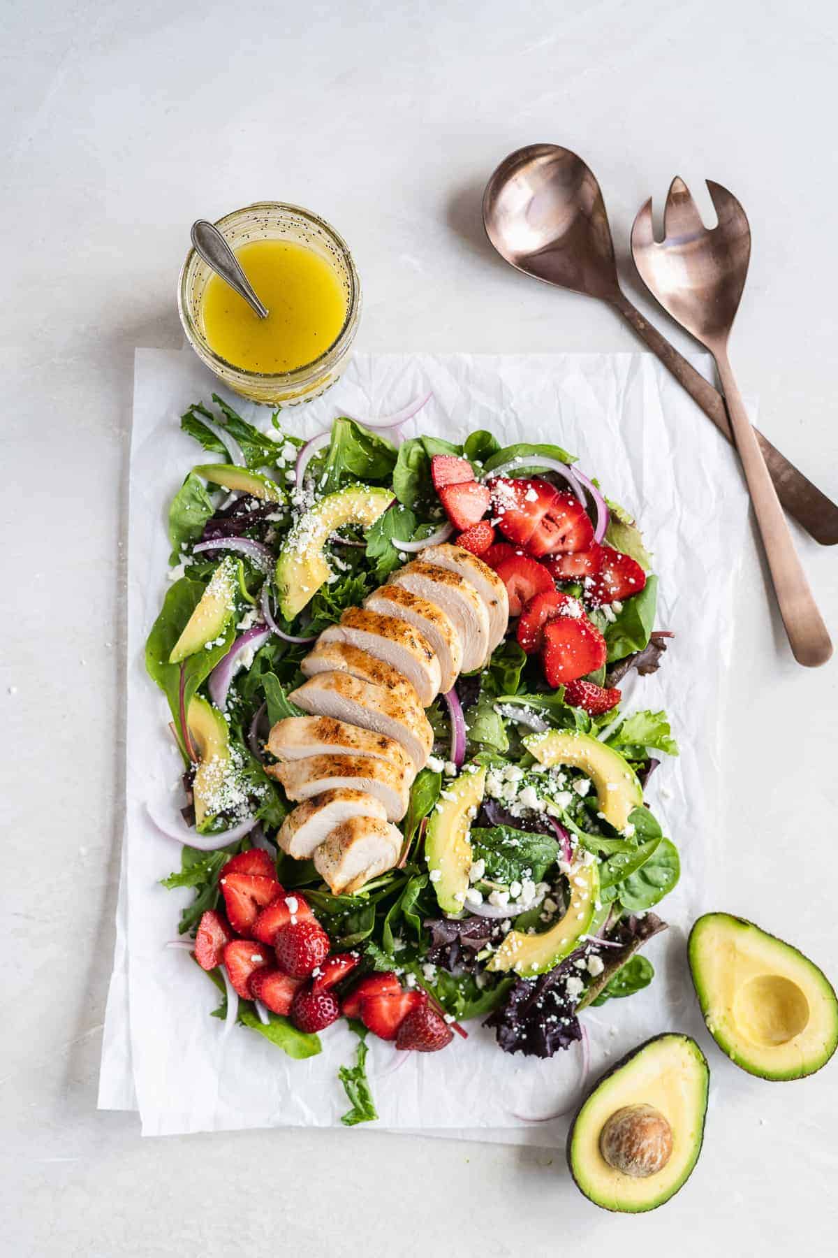 Overhead photo of the Summer Strawberry Goat Cheese Salad with Healthy Poppy Seed Dressing on white parchment paper on a marble slab.  Serving spoons and a jar of dressing are placed nearby.  