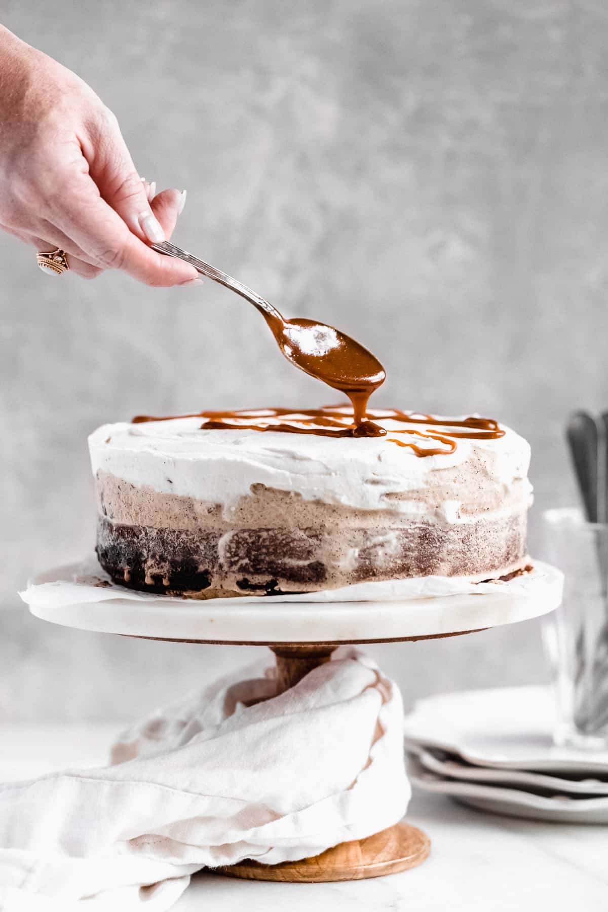 Side view photo of the Chocolate Espresso Gluten-free Ice Cream Cake sitting on a white marble cake plate.  A hand holds a spoon above that is drizzling the caramel sauce on the top of the cake.  Some small plates and utensils can be seen in the background.  