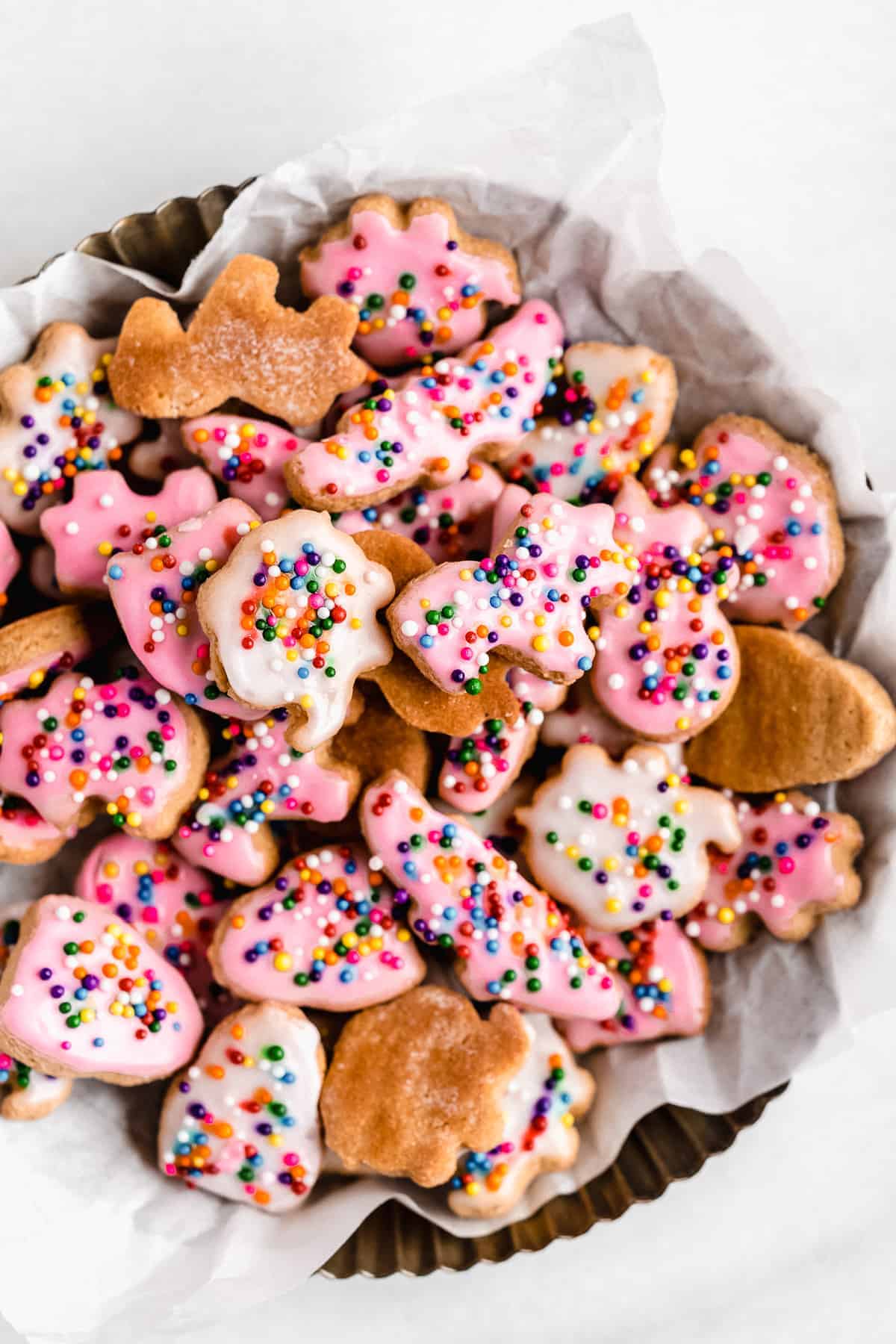 Pink and white iced animal crackers in a basket with white paper.  Rainbow sprinkles are on top of the cookies.  