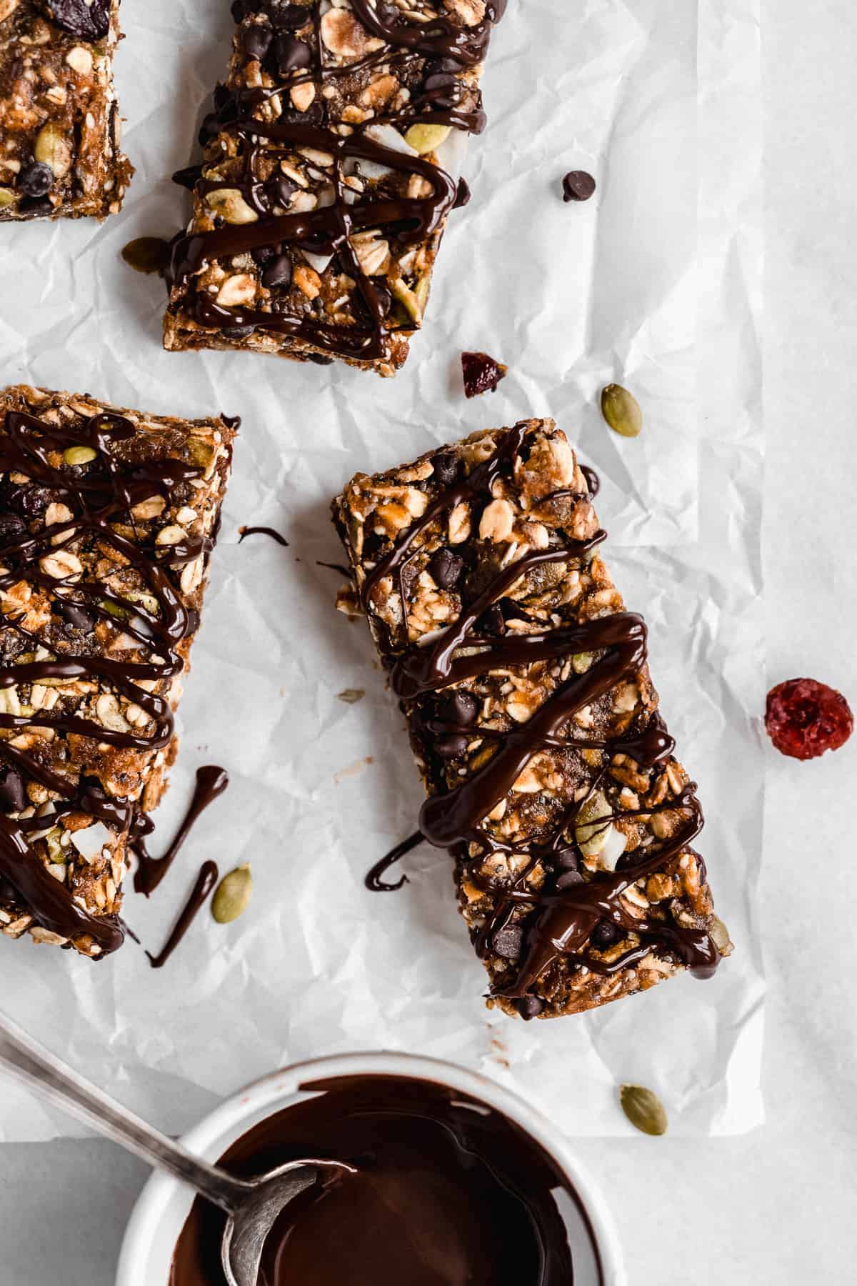 Overhead photo of several Nut-free Chocolate Granola Bars sitting on white parchment paper.  A white bowl of melted chocolate with a spoon sits nearby and has drizzled chocolate on top of the bars.  