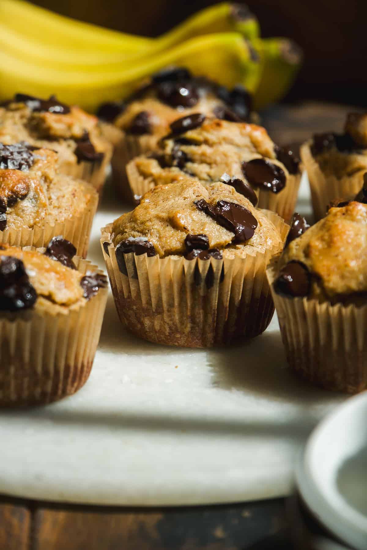 Banana chocolate chip muffins in paper liners are arranged on a white surface, with ripe bananas in the background. Sunlight highlights the golden tops and melted chocolate chips.