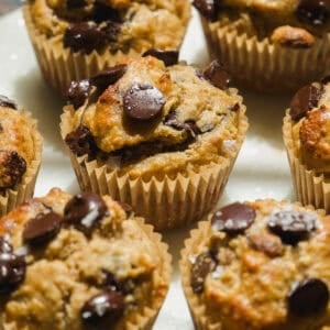 A close-up of several freshly baked muffins with chocolate chips on top, arranged closely together on a light-colored surface, with each muffin in a brown paper liner.