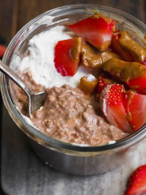 A glass jar filled with creamy chocolate overnight oats, topped with whipped cream, sliced strawberries, and a drizzle of nut butter, with a spoon tucked inside the jar.