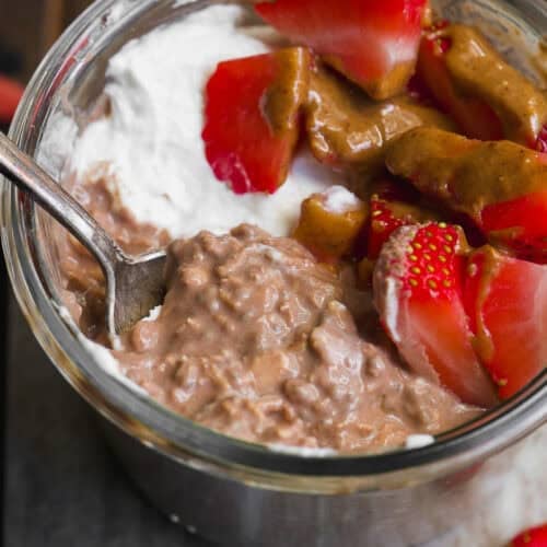 A glass jar filled with creamy chocolate overnight oats, topped with whipped cream, sliced strawberries, and a drizzle of nut butter, with a spoon tucked inside the jar.