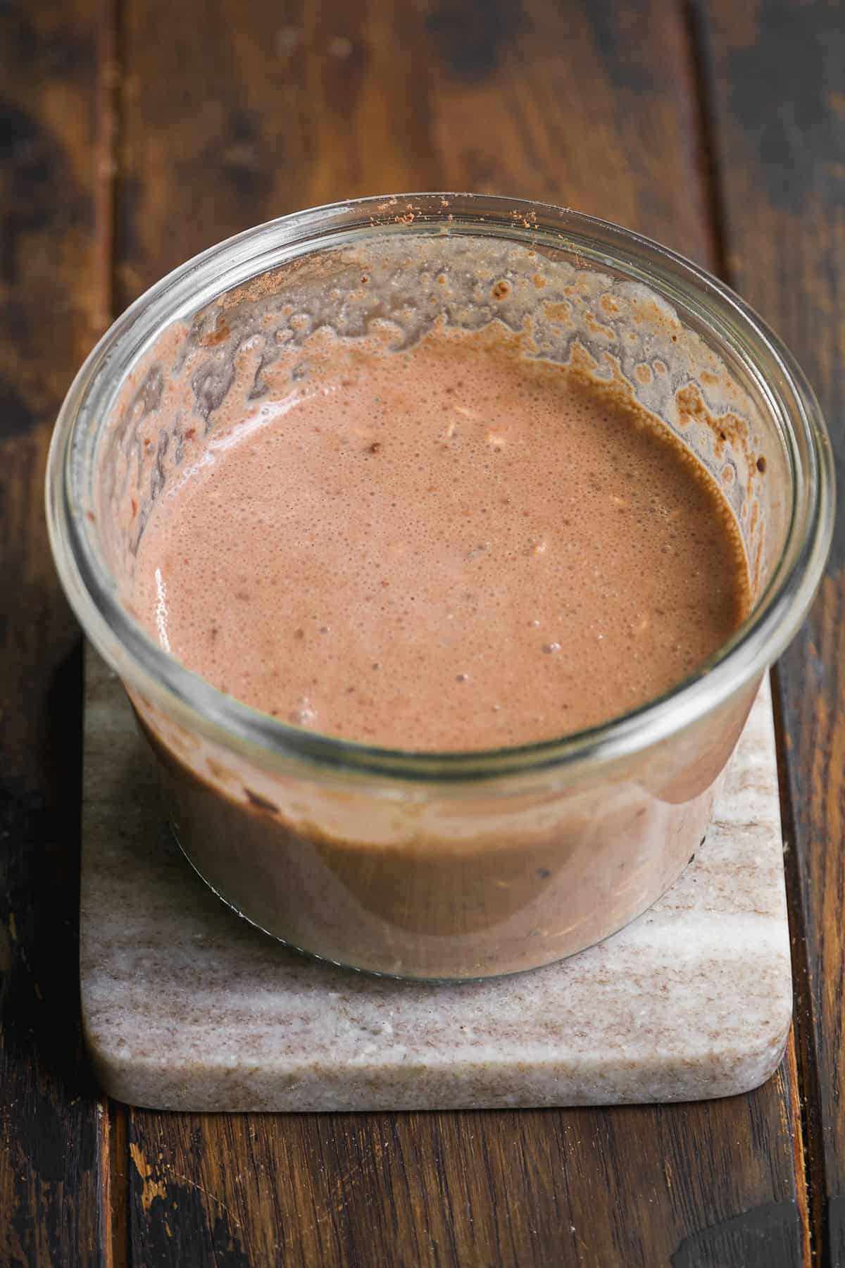 A glass bowl filled with frothy chocolate overnight oats batter sits on a square marble coaster atop a wooden surface.